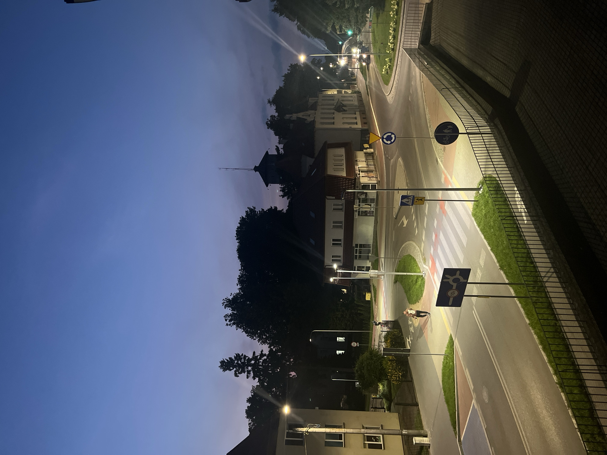 The image depicts a street scene in Ostróda, Poland, during twilight. A four-way intersection is visible, with a roundabout in the center. Several buildings, a pedestrian crossing, streetlights, and traffic signs are present. Two individuals are walking on the sidewalk, and a car is visible on the road. The sky is a deep blue, indicating the transition from day to night, and streetlights cast a warm glow on the surroundings. The overall atmosphere is calm and serene.