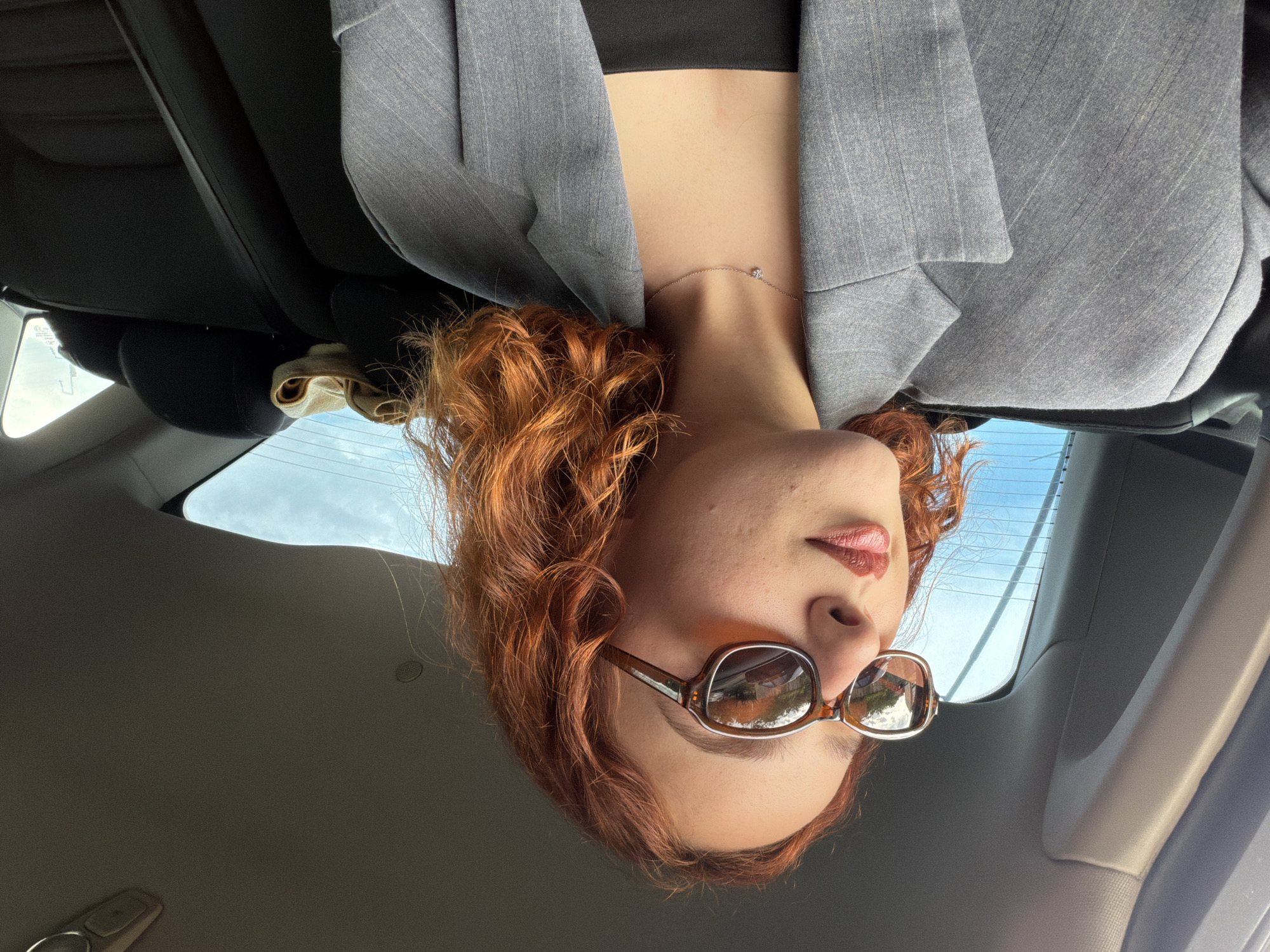 A woman with auburn, curly hair is in the driver's seat of a vehicle. She is wearing a gray blazer over a black top and a thin, delicate necklace. Sunglasses are on her face. The car interior includes black leather seats. Through the rear window, a cloudy sky can be seen. However, since this image only depicts the interior of the car, the specific location in Tbilisi, Georgia, cannot be definitively determined based on the visual data.