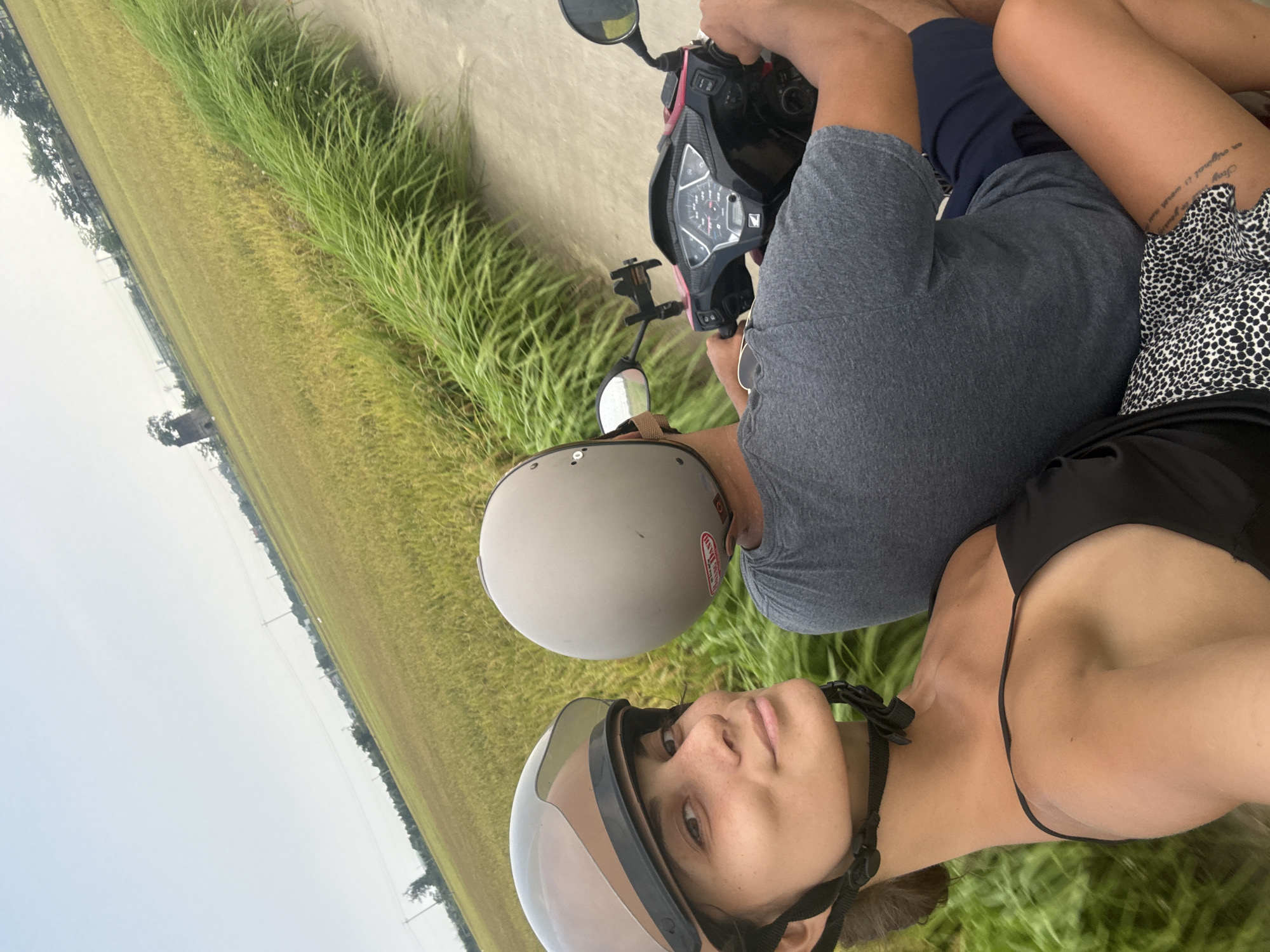 The image captures two individuals riding a scooter along a rural road, likely in Da Nang, Vietnam, as suggested by the context. The photograph is oriented vertically, giving a dynamic view of the scene.
In the foreground, a person is driving the scooter, wearing a grey t-shirt and blue shorts. Their hands are on the handlebars of a black Honda scooter, which has a visible dashboard with a speedometer and gauges, and a phone holder mounted. A grey helmet, featuring a red and white sticker with partially visible text "TEAM DASH", is worn by the driver.
Seated behind the driver is a female passenger, whose face is clearly visible as she looks directly at the camera with a neutral expression. She wears a black top and a black and white leopard print skirt or shorts. A light grey helmet with a clear visor is fastened on her head; text on the side reads "SPARTAN" with a 'D' below it. A prominent tattoo on her right thigh displays the phrases "Stay an original is correct" and "in your own way".
The scooter is moving alongside lush, expansive fields of crops, appearing green and golden-yellow, indicative of rice paddies. A narrow strip of grass borders the paved road. In the background, the landscape extends under an overcast sky, with distant trees and utility poles visible along the horizon. The soft, diffused lighting suggests it is daytime, possibly under cloudy conditions. The image conveys a sense of a leisurely ride through an agricultural landscape.