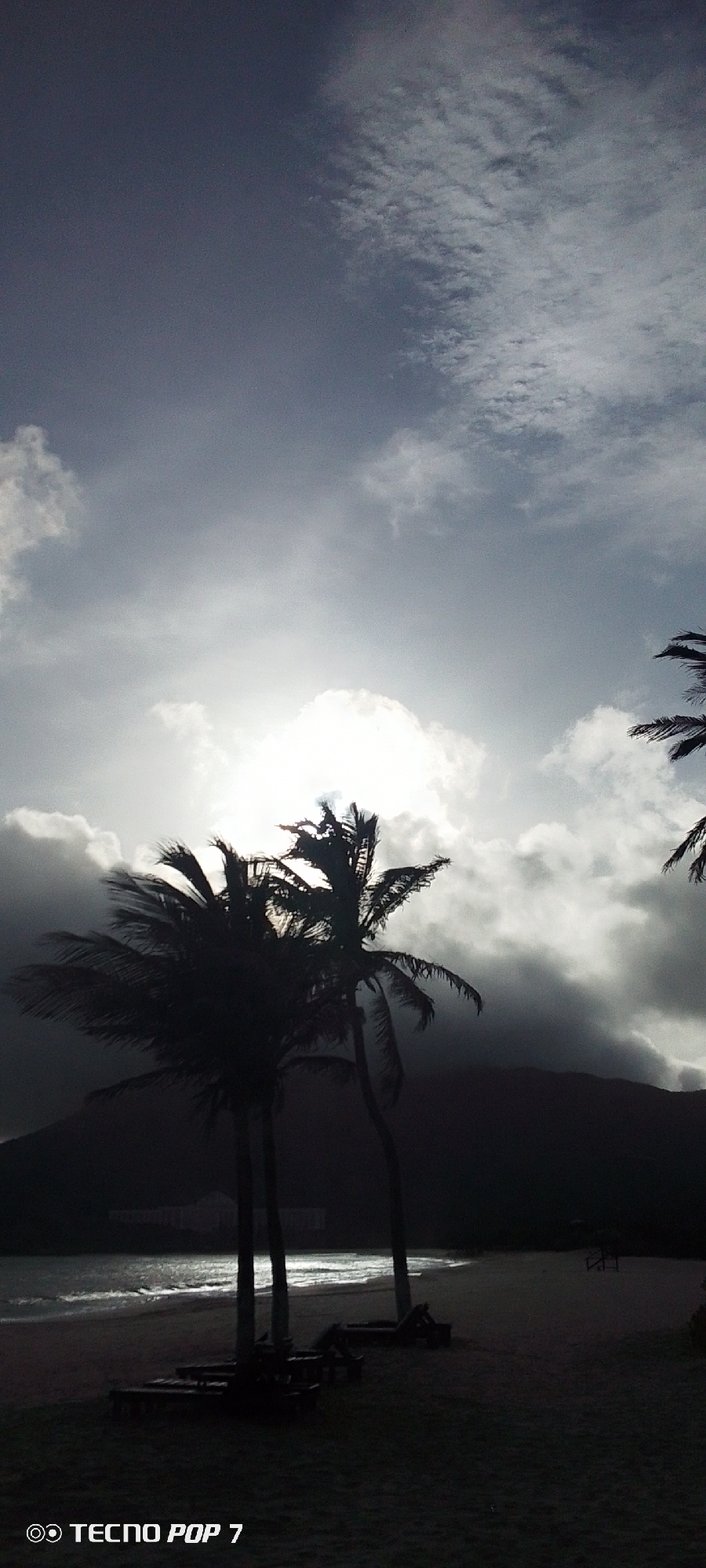 The photograph, taken with a Tecno Pop 7, depicts a beach scene in Pedro González, Venezuela. The foreground is dominated by the dark silhouettes of two palm trees, along with beach chairs and other objects. The beach sand leads to the water, where a few small waves are visible. In the background, a darkened mountain range stretches across the horizon. The sky is overcast, with heavy clouds obscuring the sun, though the sun's light still breaks through, creating a bright spot behind the palm trees.
