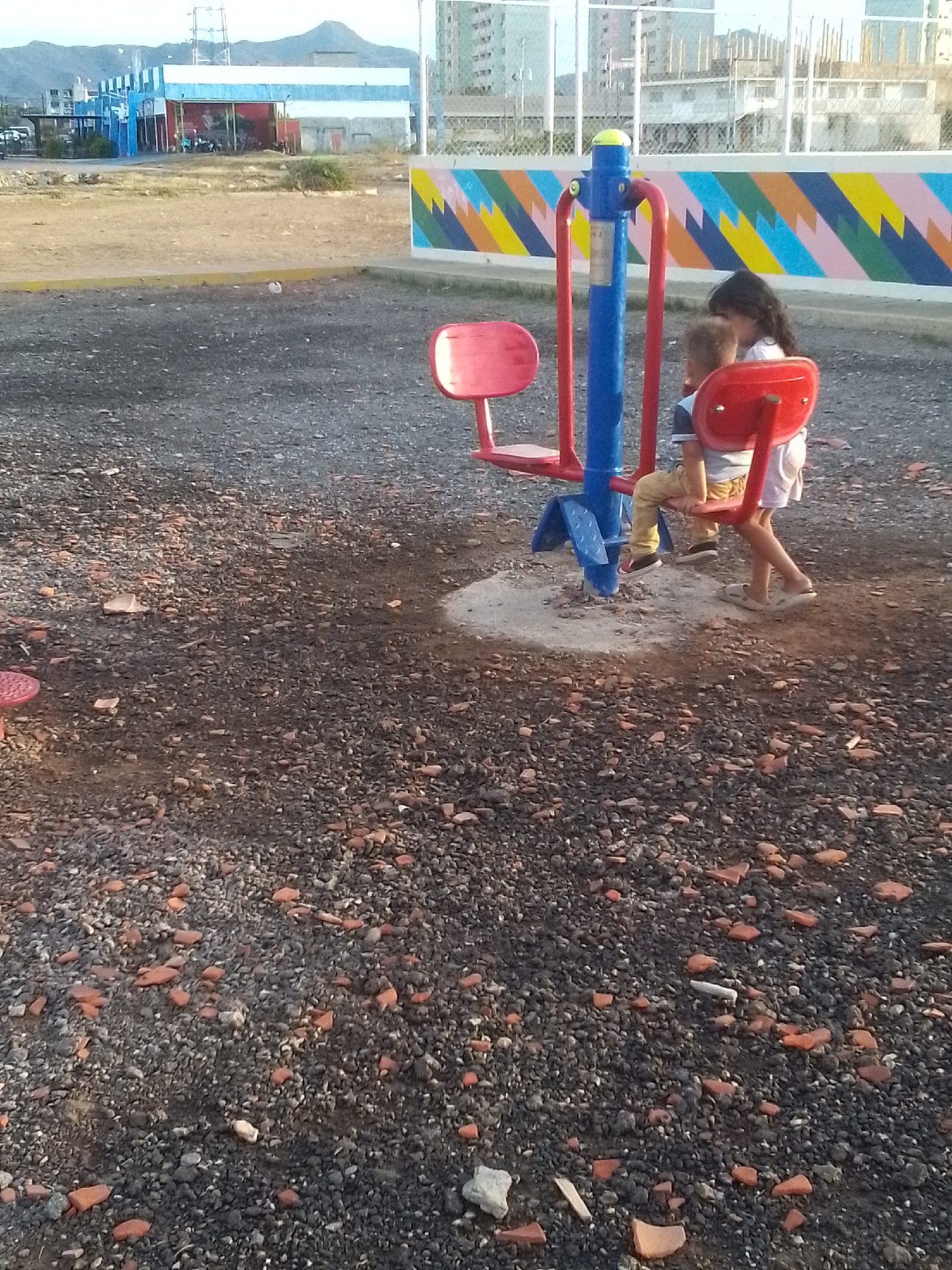 This image depicts an outdoor scene in what appears to be a recreational or open area in Juan Griego, Venezuela, during the late afternoon or early evening. The ground is covered with dark gravel mixed with numerous fragments of reddish-orange broken bricks.
Two children are the primary subjects, interacting with a piece of red and blue outdoor exercise equipment, possibly a waist twister or similar child-friendly apparatus. One child, with light brown hair and wearing a striped shirt and tan pants, is seated on one of the red seats. Behind them, a second child, with long dark hair and dressed in a white top and light-colored shorts or skirt, stands nearby, engaged in play or observation. Their faces are not clearly visible.
In the background, a low wall features a colorful geometric mural with diagonal stripes in various hues of blue, green, pink, and yellow. Beyond this wall, the urban landscape includes multi-story buildings, some appearing residential or commercial, and construction scaffolding. Further in the distance, hills or mountains are visible under a clear, warm-lit sky. To the left, a large blue and white building is also present. The overall setting suggests a public space that might be somewhat unkempt, given the ground cover. No visible text is discernible in the image.