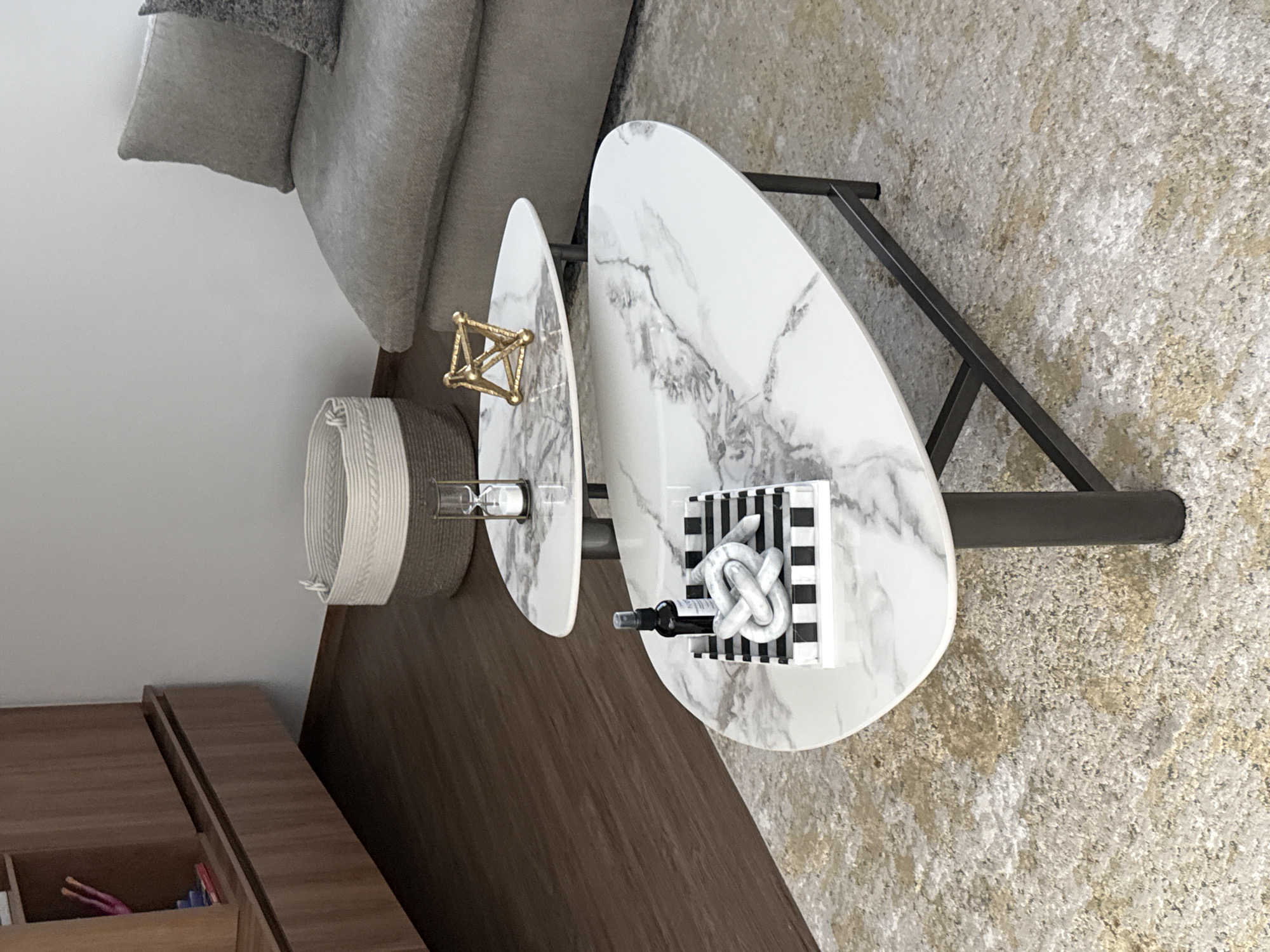 The image shows a living room interior. The angle is a high, slightly tilted, overhead view.
Dominating the scene is a two-tiered coffee table with white, marble-patterned oval tops and a dark metal frame. The table is positioned on a beige textured carpet. The table has various decorative objects. On the smaller tier, there is a gold geometric sculpture, and a small hourglass. The larger tier has a striped box with a white knotted object on top, and a small black bottle lying next to it.
To the left of the table, a section of a gray sofa is visible, including one cushion. A small woven basket sits near the sofa. Below the basket, a wood paneled cabinet runs along the left side.
The background is a plain white wall. The floor is a dark brown, possibly wood or laminate.
