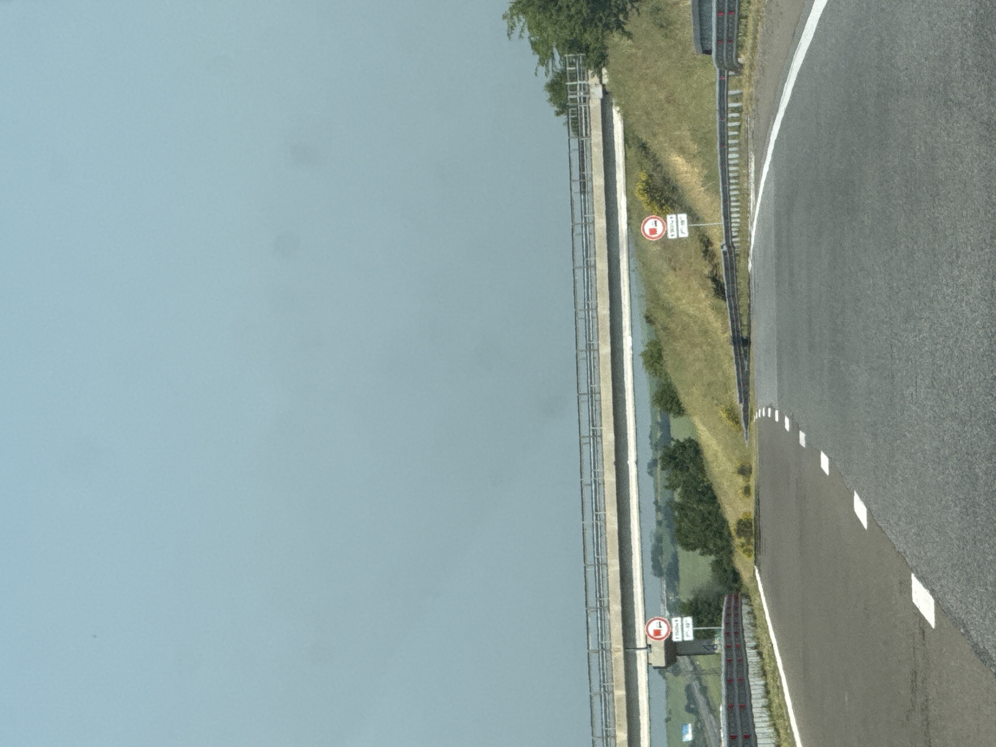 The image displays a section of an asphalt road, likely a highway, in Vratitsa, Bulgaria, under a clear, pale blue sky. A concrete overpass with metal railings traverses the frame horizontally in the mid-ground. The road surface exhibits white dashed and solid lane markings, delineating a main thoroughfare and a curving exit or entry ramp on the left. Metal guardrails are present along both sides of the road. Beneath a structural support of the overpass on the left, a circular regulatory sign with a red border and black truck icon indicates a restriction against trucks, supplemented by a rectangular sign displaying "1500m" and "7h-19h". An identical truck restriction sign, also accompanied by a rectangular sign partially visible with "7h-19h", stands on the right grassy embankment. The environment consists of green, grassy slopes rising on either side of the road, interspersed with trees and bushes. A faint white vehicle is visible in the far distance on the left side of the road. No individuals or active vehicles are present in the foreground or mid-ground.