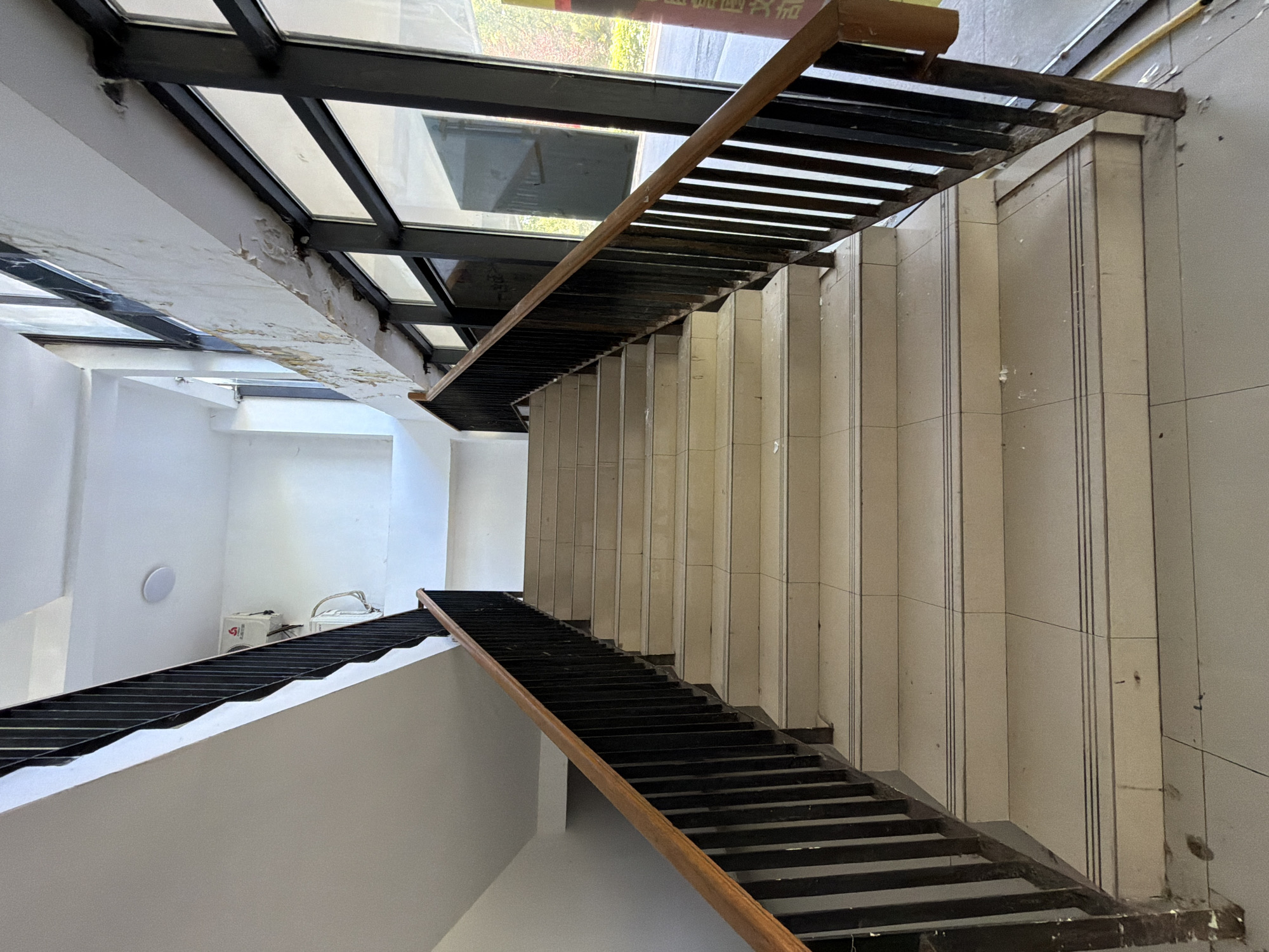 The image shows an interior staircase with cream-colored tiled steps and black metal railings with a wooden handrail. The staircase leads upwards. To the right of the staircase, there are large windows with dark frames, offering a view of the exterior, which appears to be a street or outdoor area with some greenery visible. The walls are painted white. There are some signs of wear and tear, particularly on the ceiling area above the windows, where the paint is peeling. An air conditioning unit is visible mounted on the wall near the top of the staircase. The scene depicts a part of the Jiangjin district in China, likely an entrance or common area in a building. No people are present in the image.
