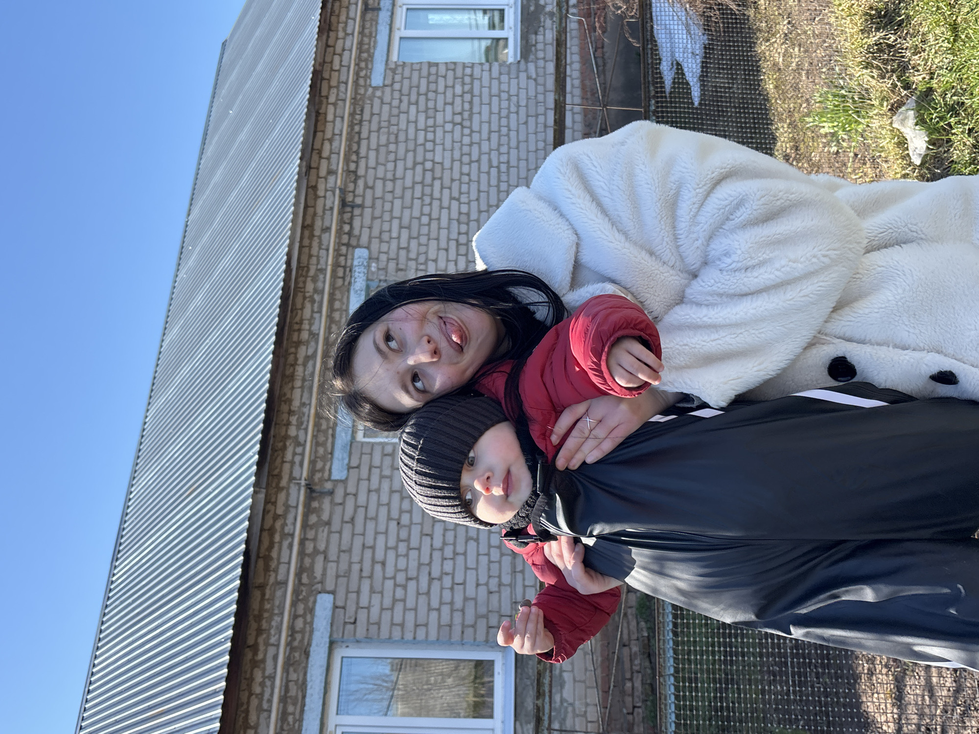 Two individuals are positioned outdoors against the backdrop of a two-story building in Фоки, Russia. The upper part of the building features a corrugated metal roof, while the lower portion is constructed of brick, containing at least one window. One individual, an adult female, is wearing a white furry coat and a red jacket. She is embracing the other individual, a child wearing a dark knit cap, a black and white tracksuit, and a red jacket. The woman is making a playful facial expression, sticking out her tongue and looking upward.