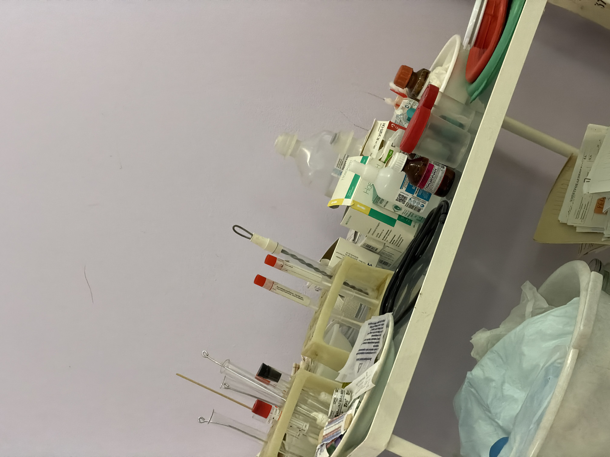 The image displays a cluttered medical preparation area situated within a facility in Chaikovsky, Russia. A white, two-tiered utility cart is positioned against a light lavender wall, holding various medical and laboratory supplies. On the upper shelf, multiple clear glass test tubes, some containing red-stoppered or red-capped sample tubes (e.g., Vacuette brand), are secured in beige plastic racks. Accompanying these are wooden applicators and metal stirring rods. Several bottles and boxed medications are present, including those labeled in Cyrillic as "Натрия хлорид" (Sodium Chloride), "НО-ШПА" (No-Spa), and "ГЛИЦЕРИН" (Glycerin). Also on this shelf are clear plastic specimen containers with red lids, brown glass bottles, a white rectangular basin, and distinct red and green plastic trays. The lower shelf contains a white bucket filled with crumpled light blue disposable medical material, and a stack of documents, one partially visible and labeled "ЭЛЕКТРОКАРДИОГРАФИЯ" (Electrocardiography). No individuals are visible in the scene, which appears to be a functional, albeit disarranged, diagnostic or treatment staging area.