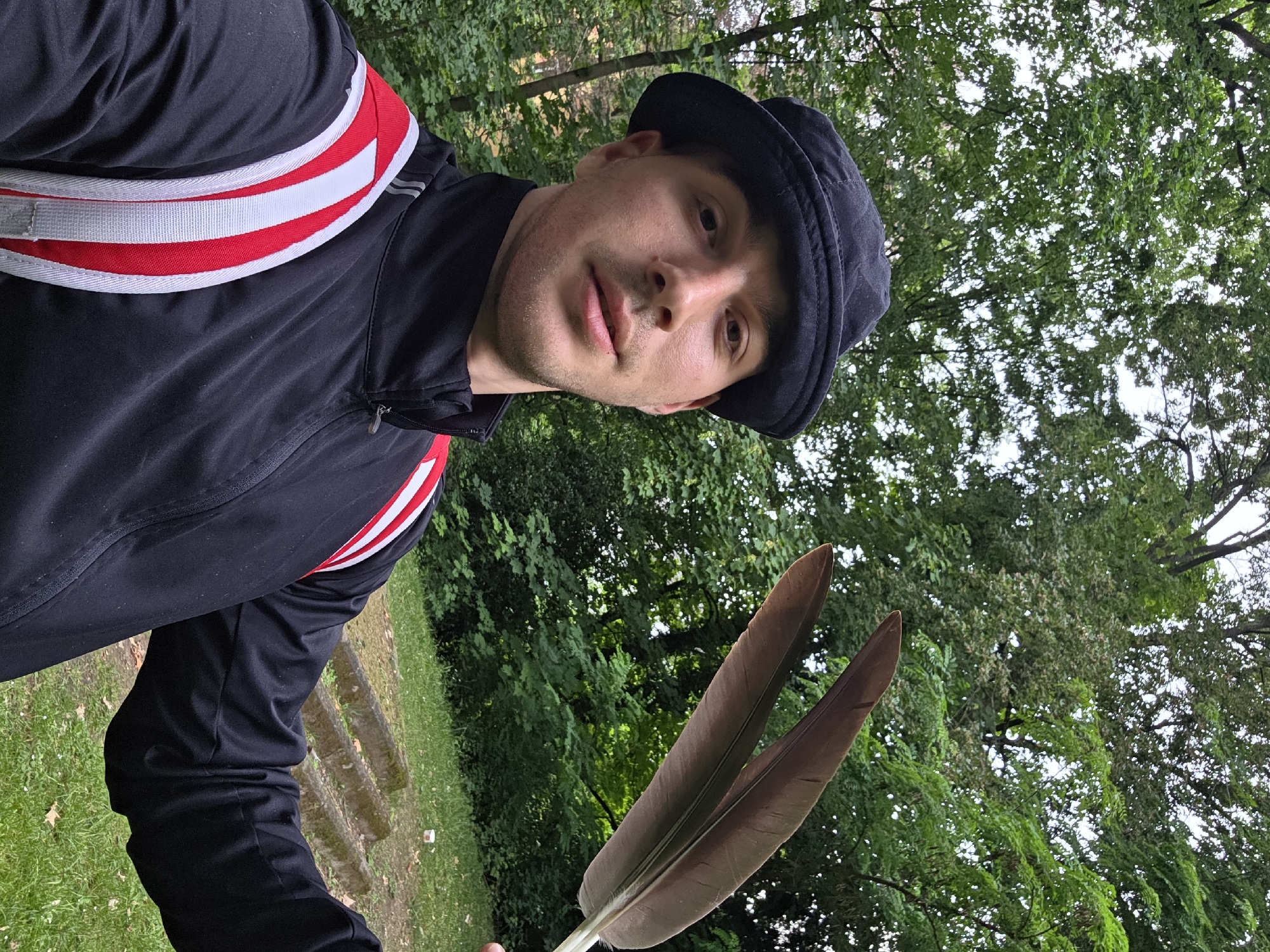 In an outdoor, tree-filled environment, a young Caucasian male is holding two large brown feathers in his right hand. He is wearing a black bucket hat, a black zip-up jacket with red and white stripes on the shoulders, and a backpack with similar red and white straps. The man is looking at the camera, and the background consists of green trees and a grassy area with concrete steps. The image appears to be a selfie.
