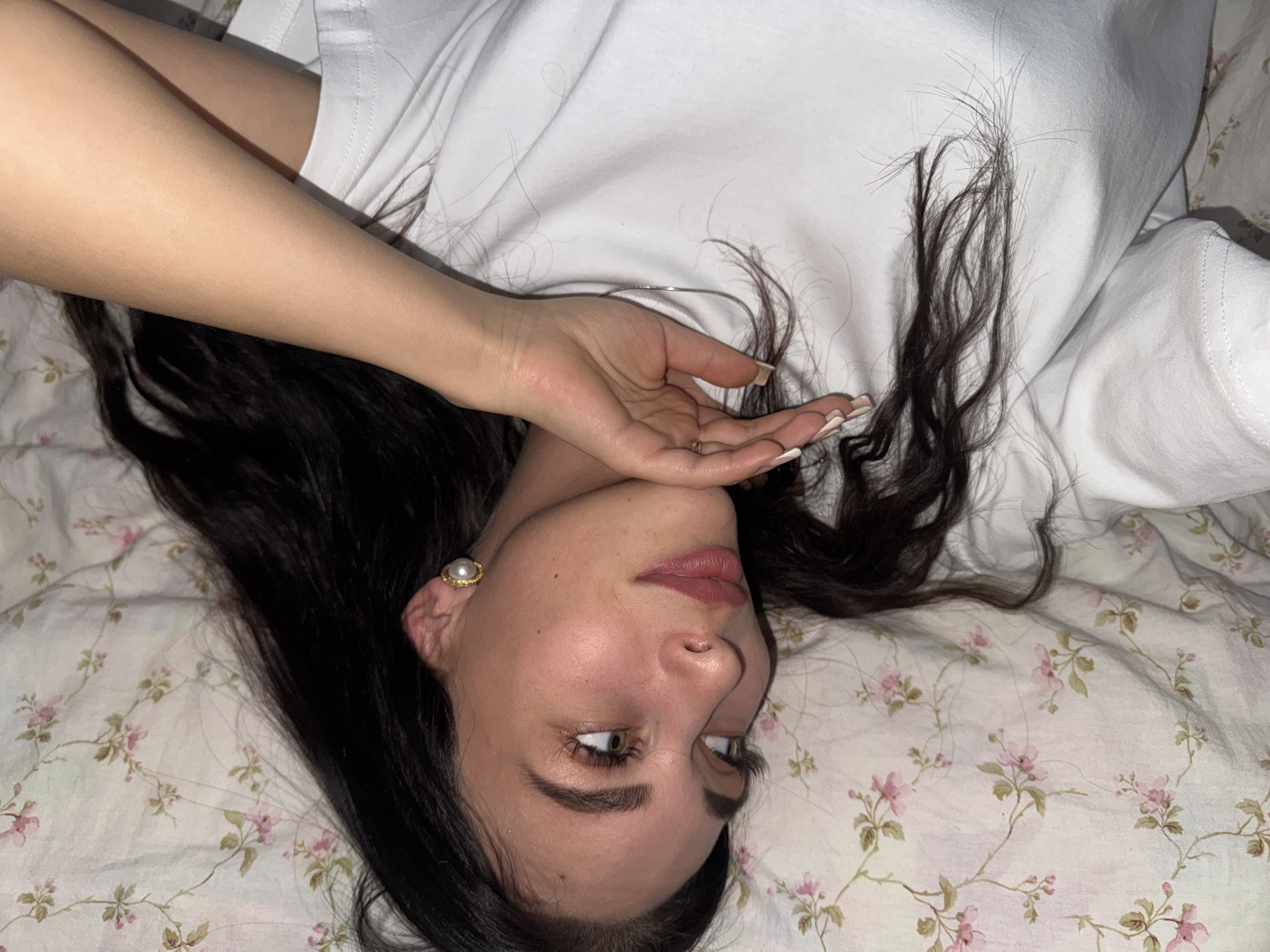 An angled, close-up shot captures a young woman lying on a bed, potentially in Chaikovsky, Russia. Her head is tilted, bringing her face into the lower right of the frame, while her body extends upwards to the left. She has long, dark hair, some of which is spread out on the floral-patterned bedding and behind her head. She is wearing a simple white t-shirt.
Her right hand (from the viewer's perspective) is raised, fingers gently curled, partially obscuring her chin and mouth, though her subtly parted, pink-tinted lips are visible. Her nails are manicured with light-colored polish and French tips, and a ring adorns her ring finger. A distinctive pearl earring with a gold-toned trim is visible in her left ear. Her eyes, appearing light-colored, are open and gazing upwards and to her left, creating a somewhat introspective or engaging expression. Her eyebrows are well-defined, and she has visible eyelashes.
The bedding beneath her features a delicate pattern of pink flowers with green leaves and thin stems on a light, possibly white or off-white, background. A plain white fabric is also visible, likely a pillow or another part of the bedding, beneath her upper body. The soft, indoor lighting contributes to a calm and intimate atmosphere. There is no visible text in the image.