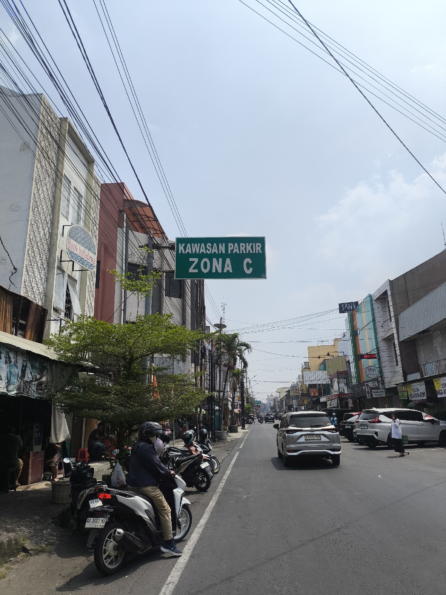 The image depicts a street scene in Surakarta, Indonesia, likely a commercial area based on the presence of shops and signage. A green sign overhead reads "KAWASAN PARKIR ZONA C," indicating a parking zone. The street is paved and appears to be a two-way road with vehicles moving in both directions. Several motorbikes are parked along the left side of the street, some with riders, suggesting a busy area. On the right side of the road, cars are also parked, including a silver SUV and two white SUVs. Pedestrians are visible, including a person walking across the street on the right side. The buildings lining the street are of varying architectural styles and colors, with some featuring awnings and signs for businesses. Power lines are strung across the sky. The sky is mostly clear with a few wispy clouds. The overall impression is of a typical urban street in Indonesia on a sunny day.