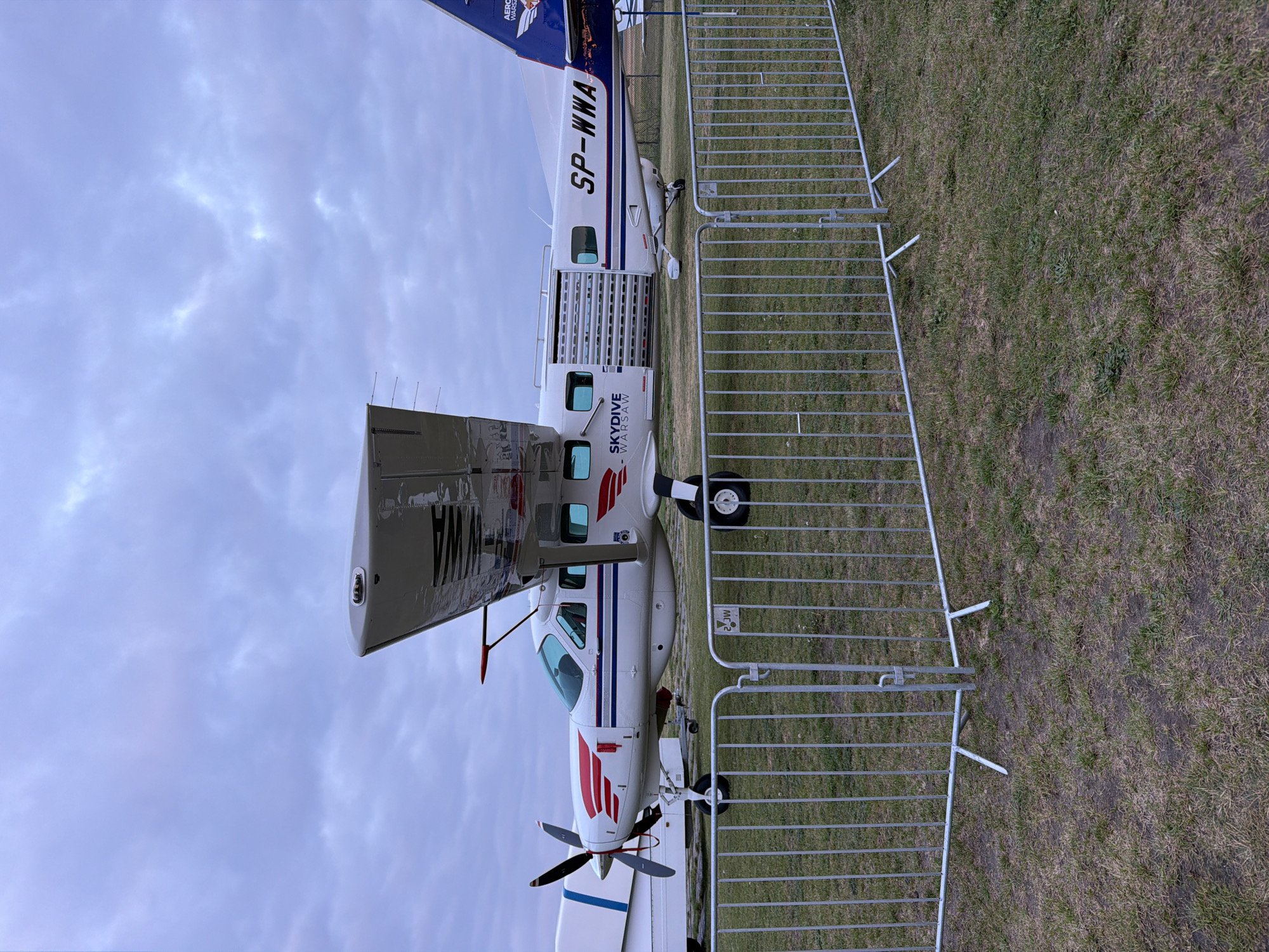 The image captures a side view of a small, white airplane bearing the registration code "SP-WWA" on its tail. The plane is marked with the words "SKYDIVE WARSAW" in large letters. It has a red stripe design on the front and tail. The rear door is open, revealing a caged interior. A temporary metal fence is erected alongside the plane, with a grassy area visible on both sides. The sky is overcast. The location is unspecified.