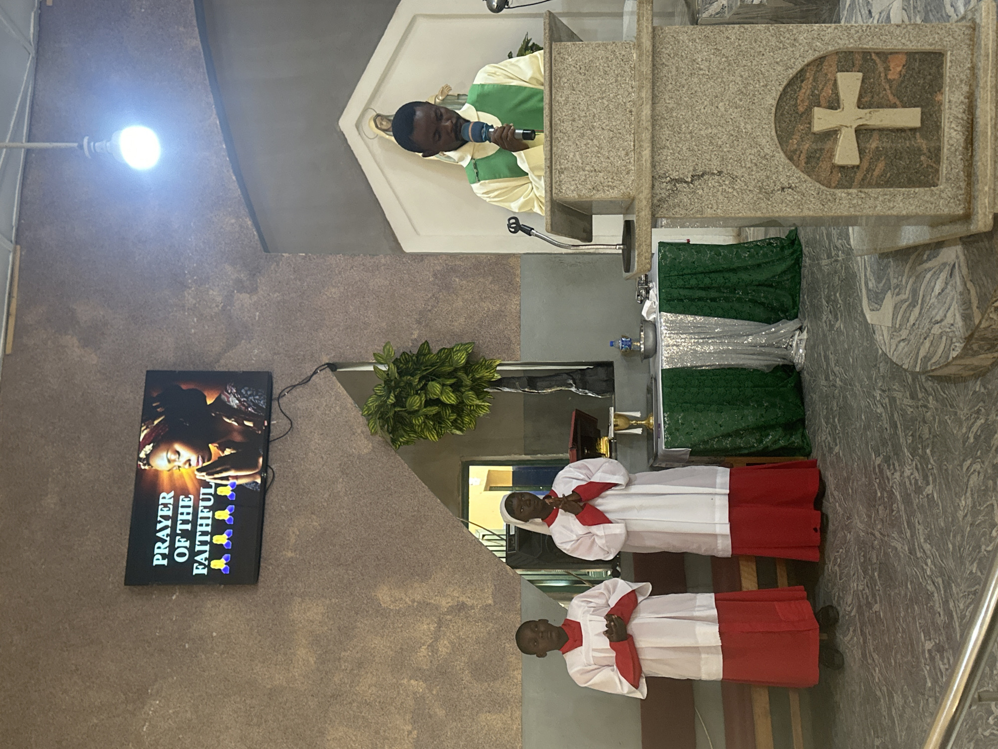 In Akwanga, Nigeria, a religious service is taking place in what appears to be a church. Two young people stand attentively on the left, dressed in white robes with red accents, their hands clasped in prayer. To the right, a clergyman, also in religious attire, stands behind a stone podium, speaking into a microphone. Above them, a television screen displays the text "PRAYER OF THE FAITHFUL" alongside an image of a woman in prayer and several yellow praying hands icons. The environment is decorated with green and silver draped fabric on a table in the background, and a large potted plant is visible on the right. The lighting appears to be artificial, with a bright bulb hanging from the ceiling.