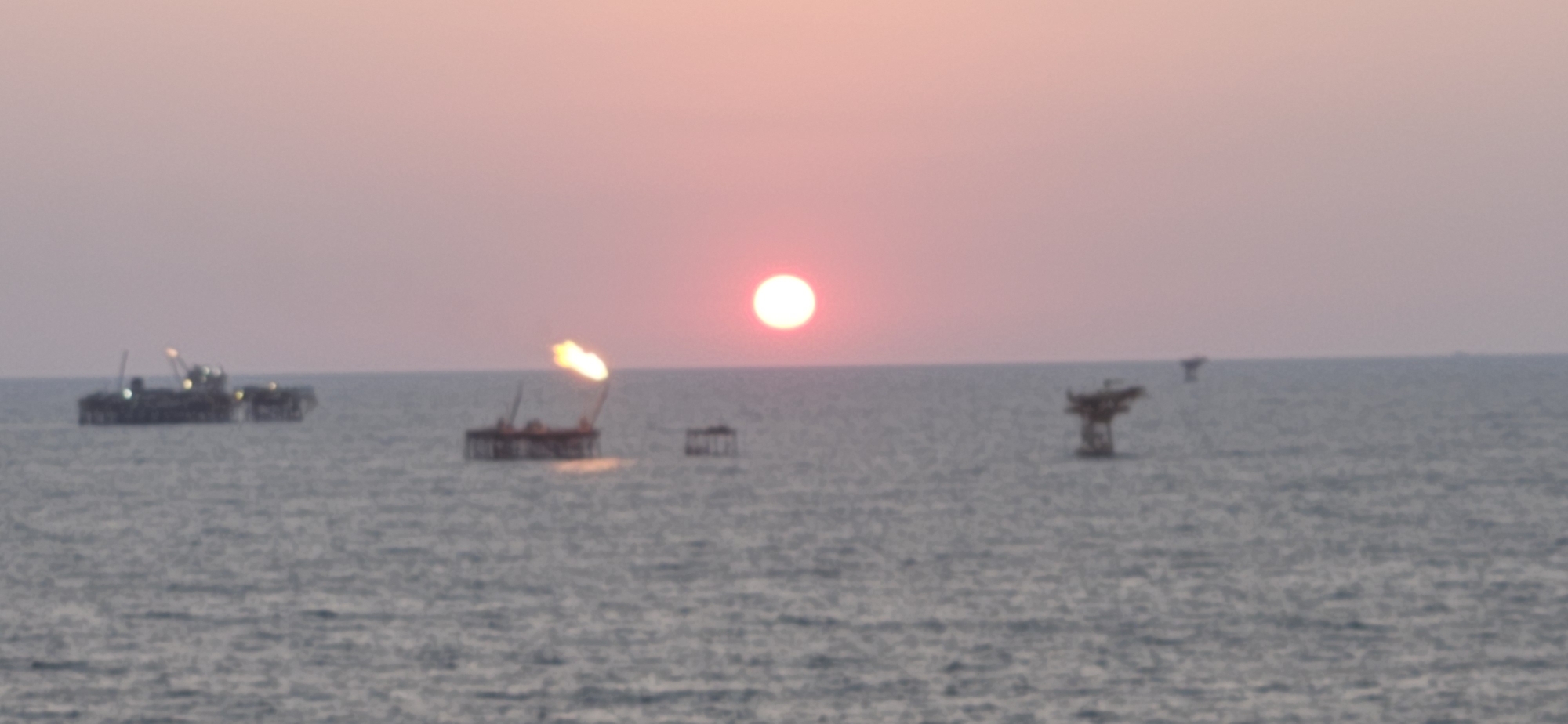 This is a wide shot photograph of an offshore oil rig at sunset. The sun is a bright, round orb positioned in the upper center of the frame, casting a soft pinkish-orange glow across the sky and reflecting on the water. The sea is a calm, deep blue with gentle ripples. In the foreground, on the left side of the image, is a large oil rig structure with multiple platforms and lights. A flame is visible on the rig, suggesting active operation. Further in the distance, other smaller structures, possibly other rigs or platforms, are scattered across the horizon. The overall atmosphere is serene and industrial, with the natural beauty of the sunset contrasting with the man-made structures. The location is the sea, offshore.
