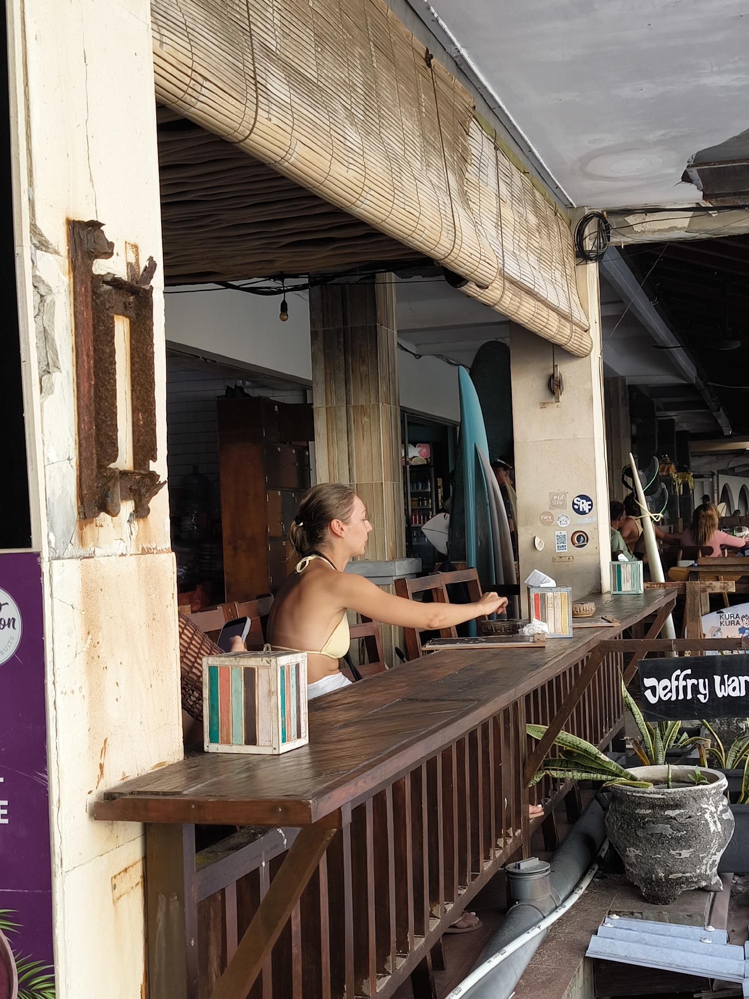 The image depicts an outdoor dining area, likely in Uluwatu, Indonesia, based on the architectural style and signage. A woman with blonde hair tied back, wearing a light yellow bikini top, is seated at a wooden bar counter. She is reaching towards something on the counter.
Behind her, surfboards are propped up against a wall adorned with stickers. Other patrons are visible in the background. The bar counter has decorative wooden blocks and a planter containing leafy plants at one end. The overall atmosphere suggests a casual, beachside establishment.