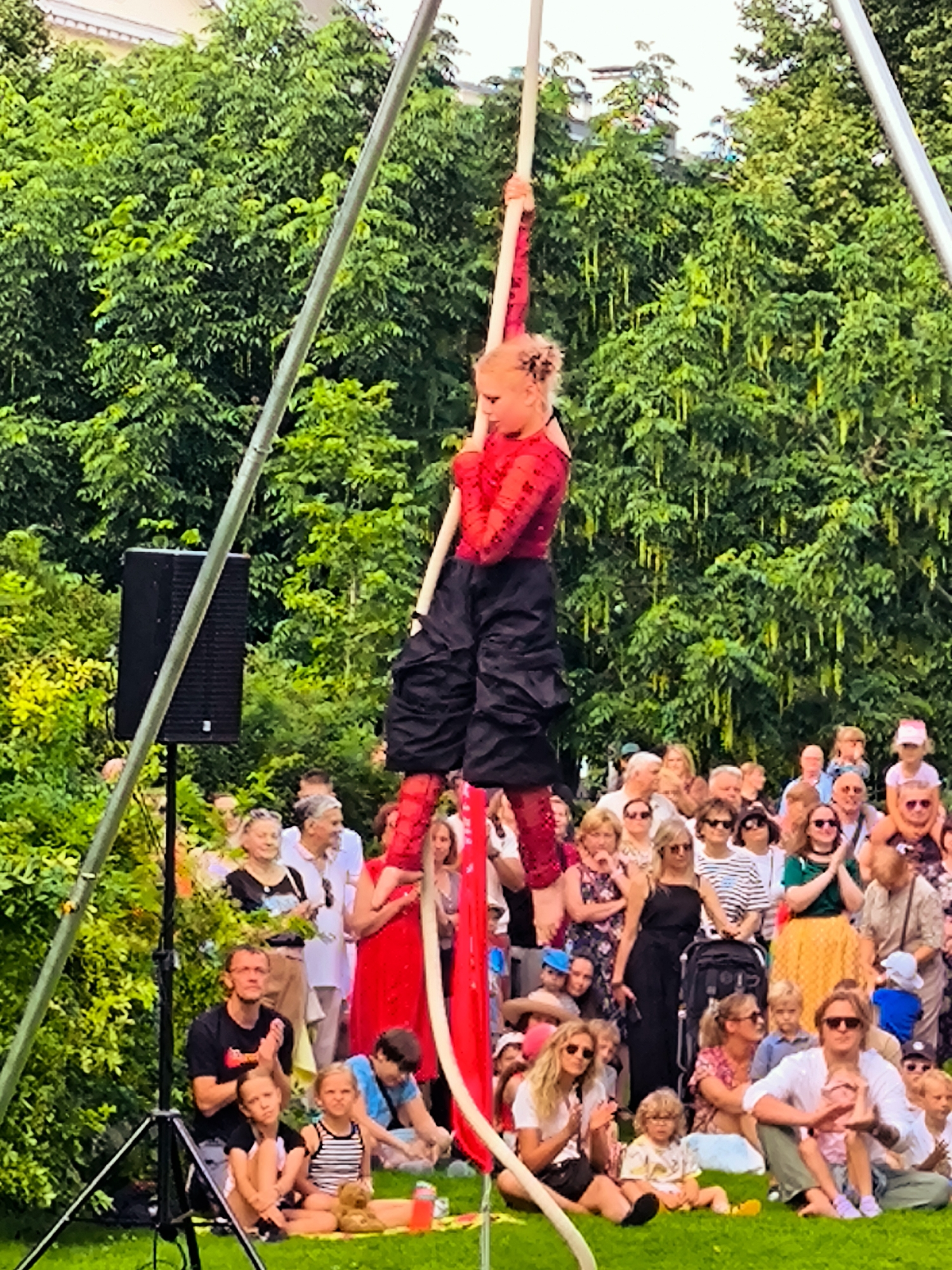The image captures an outdoor performance in Riga, Latvia, likely during the daytime under clear weather conditions, given the bright lighting and vibrant greens of the surrounding trees. A young performer, a girl with blonde hair, is suspended mid-air, skillfully balancing on a tall, vertical pole. She is dressed in a striking red, long-sleeved top adorned with glitter and voluminous black pants, with red accents on her legs.
Beneath the performer, a crowd of spectators is gathered on a lush green lawn, watching the show. The audience comprises people of all ages, many sitting on the grass, while others stand, creating a lively and engaged atmosphere. Among the audience members, some are clapping and observing the performance with visible interest. A speaker system is visible to the left of the performer, suggesting amplification for the event. The overall scene is one of community enjoyment and appreciation for a performance, set against a backdrop of dense, green foliage.