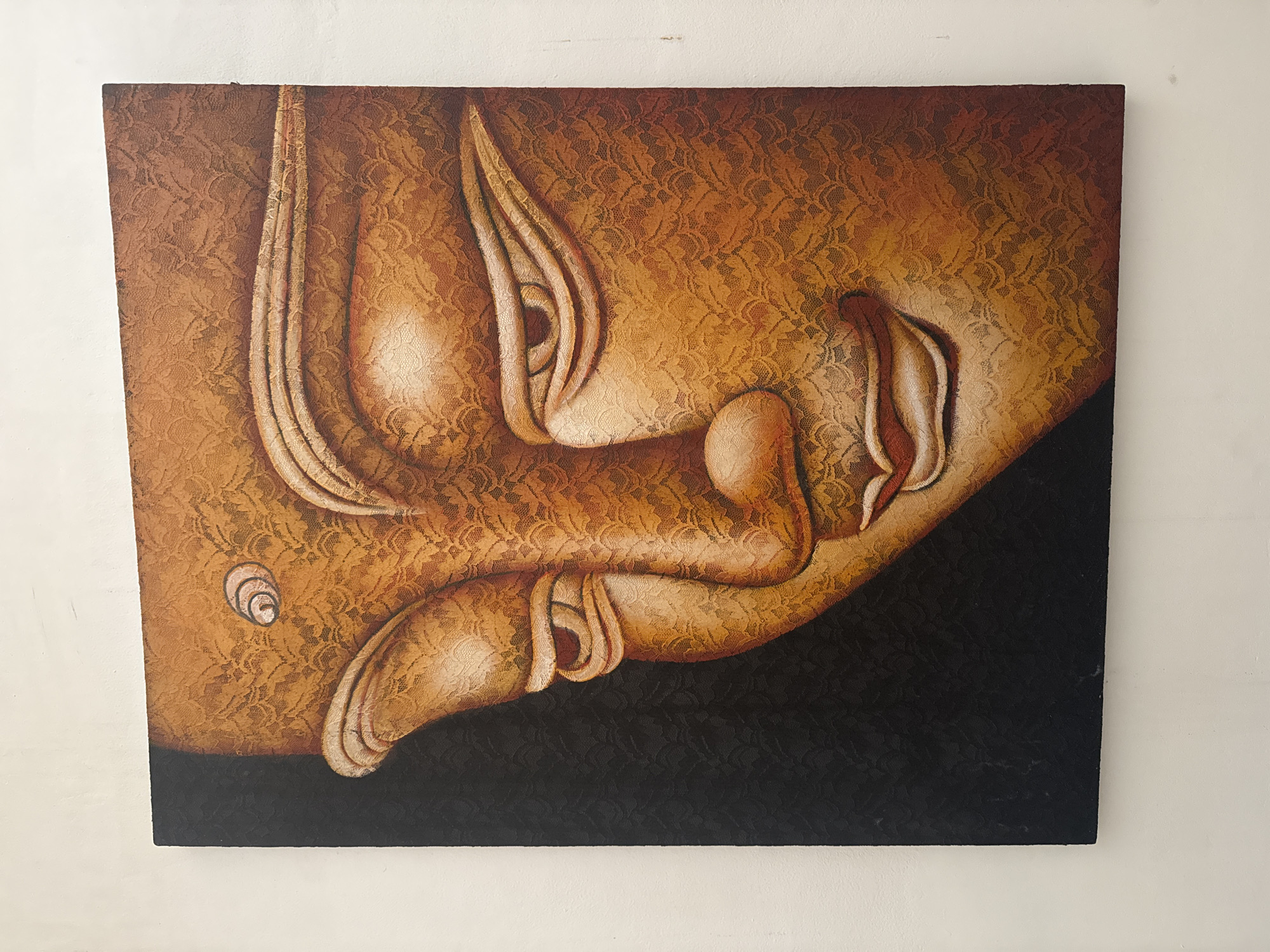 The image shows an oil painting of a reclining Buddha head against a dark background. The head is adorned with an ornate lace pattern. The picture hangs on a white wall, with the composition tightly cropped to focus solely on the artwork. The painting was likely taken in North Kuta, Indonesia.