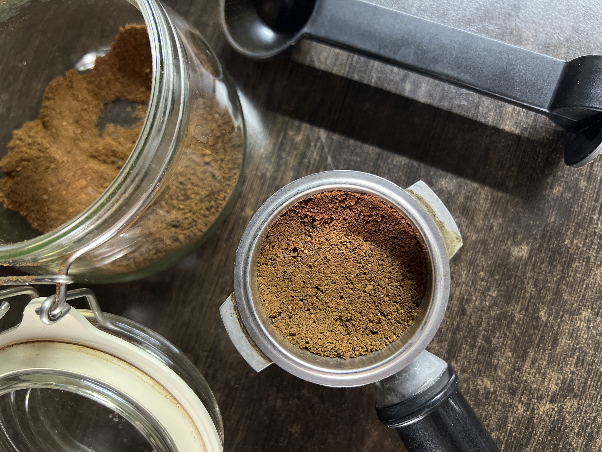 A close-up, overhead shot shows a portafilter filled with finely ground coffee, sitting on a dark wooden surface. To the right of the portafilter, a black coffee scoop lies at an angle. In the background, a glass jar with a clamp lid contains more ground coffee. The lighting casts shadows across the surface, with the lid of the jar slightly open and a hint of its rubber seal visible.