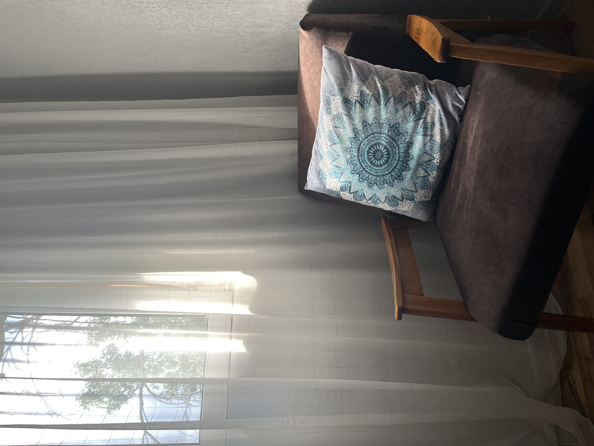 The image depicts a brown armchair with a decorative blue and white patterned pillow. The armchair is positioned next to a window covered with sheer white curtains. Sunlight streams through the window and curtains, casting shadows and illuminating the scene. The background consists of a textured wall on the right and a glimpse of greenery outside the window. The overall impression is one of a quiet, indoor space. This scene could be any domestic interior in Tashkent, Uzbekistan, as there are no identifying features to place it in a specific location within the city.