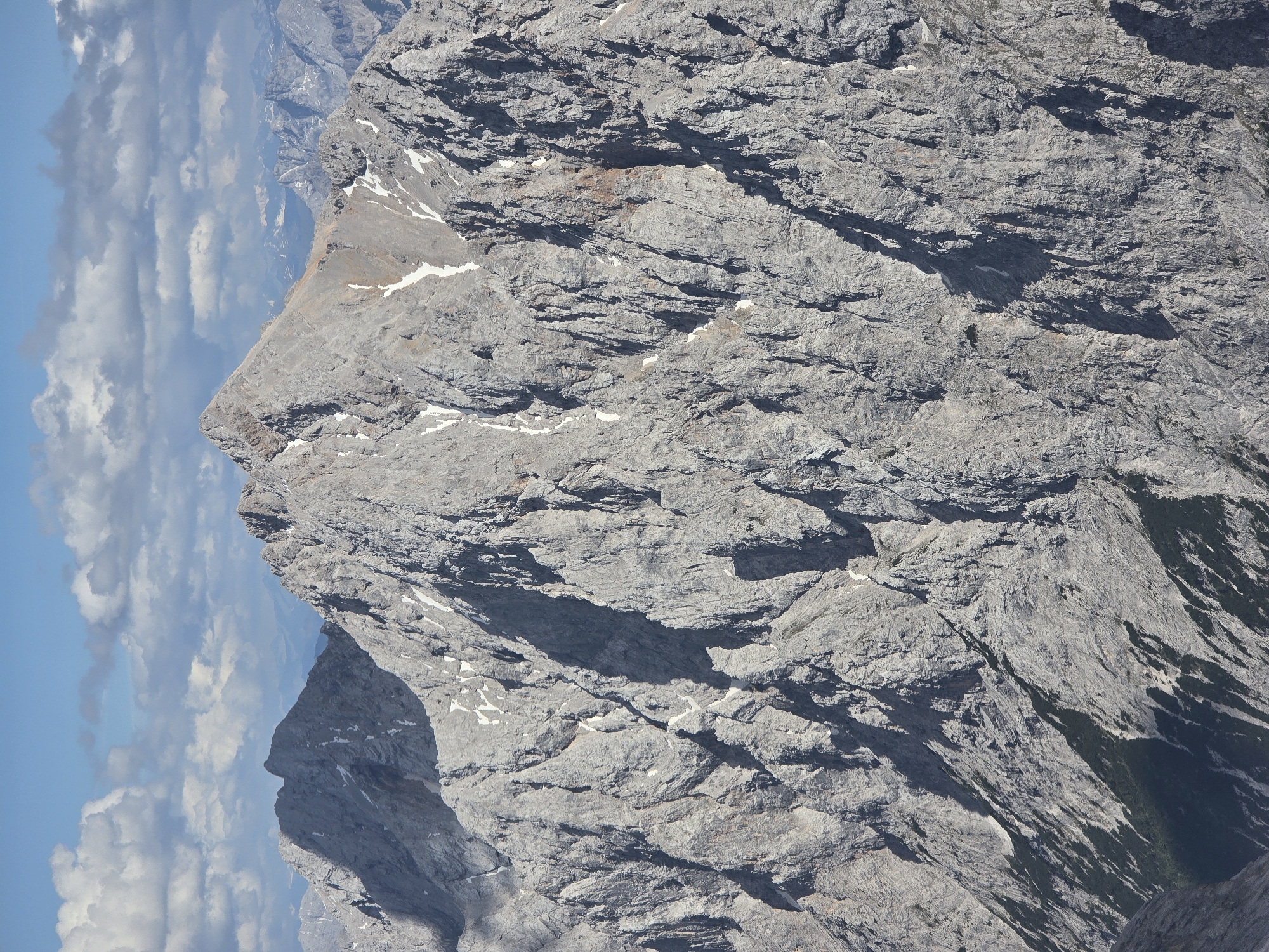 The image depicts a close-up, aerial view of a rugged mountain range under a partly cloudy sky. The mountains are predominantly gray rock with visible textures and crevices, indicating erosion and geological formations. Patches of snow are scattered across the slopes, suggesting a high altitude or a cold season. The foreground shows steep, rocky terrain, with some darker areas that could be shadows or areas of vegetation lower down the slopes. In the distance, other mountain peaks are visible, fading into a hazy blue, suggesting a vast mountain landscape. The sky above is a vibrant blue, with scattered white cumulus clouds.
This image is likely of the Bavarian Alps, in the vicinity of Garmisch-Partenkirchen, Germany. The prominent mountain in the center is likely the Zugspitze, Germany's highest peak, or one of its surrounding formidable peaks. Garmisch-Partenkirchen is a well-known alpine resort town at the foot of the Zugspitze massif, offering panoramic views of these very mountains. There are no people, animals, or man-made structures visible in this specific image.