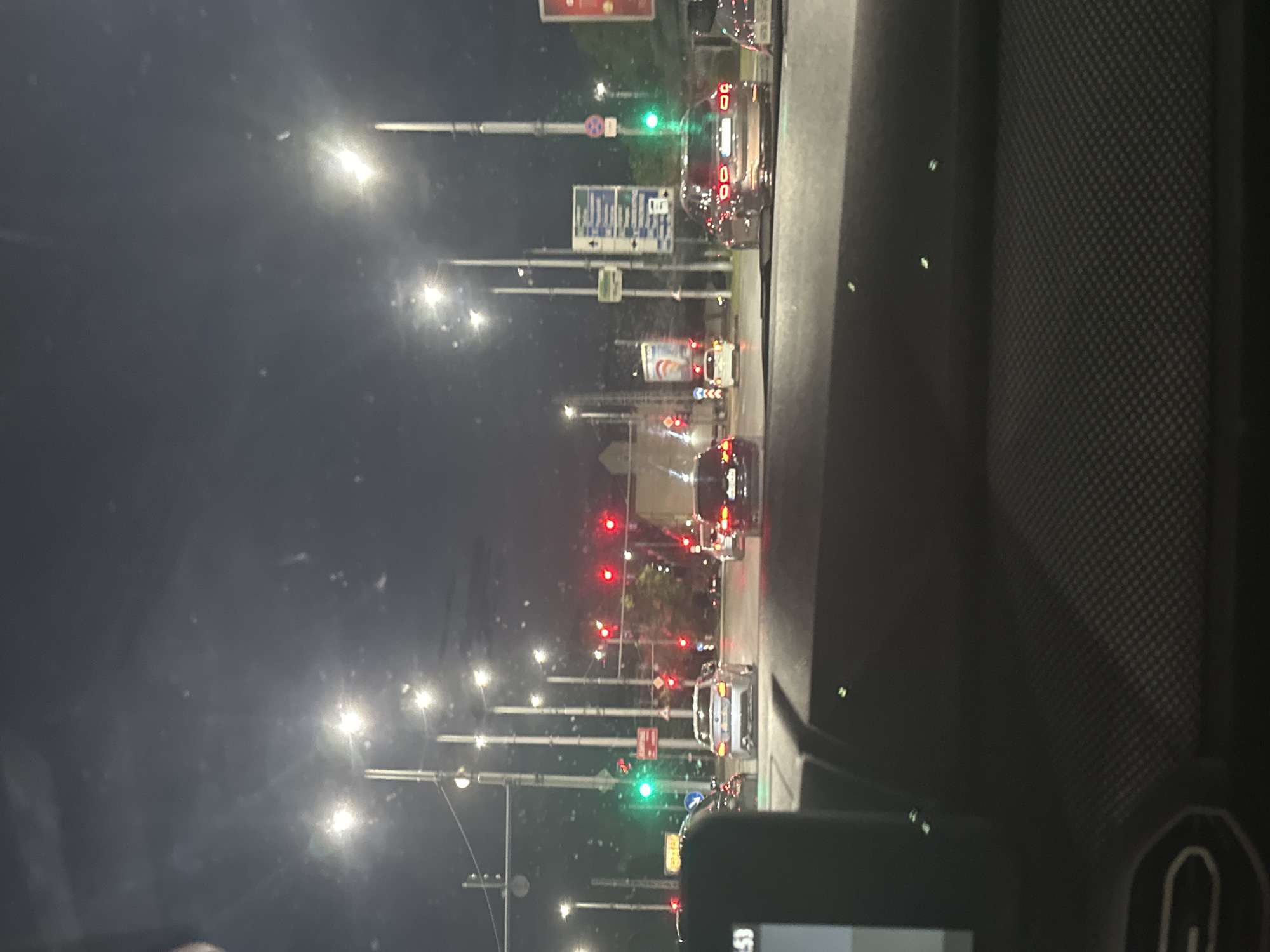 The image depicts a street scene at night in Plovdiv, Bulgaria, likely at an intersection with traffic lights. The perspective is from inside a vehicle, looking forward through the windshield. Several cars are visible on the road, some with their headlights and taillights illuminated. Streetlights line the road, casting a glow on the pavement and surrounding buildings. Traffic lights are prominently displayed, with some showing red and one showing green. Road signs are also visible, providing directional information. The overall atmosphere is one of urban nighttime traffic.