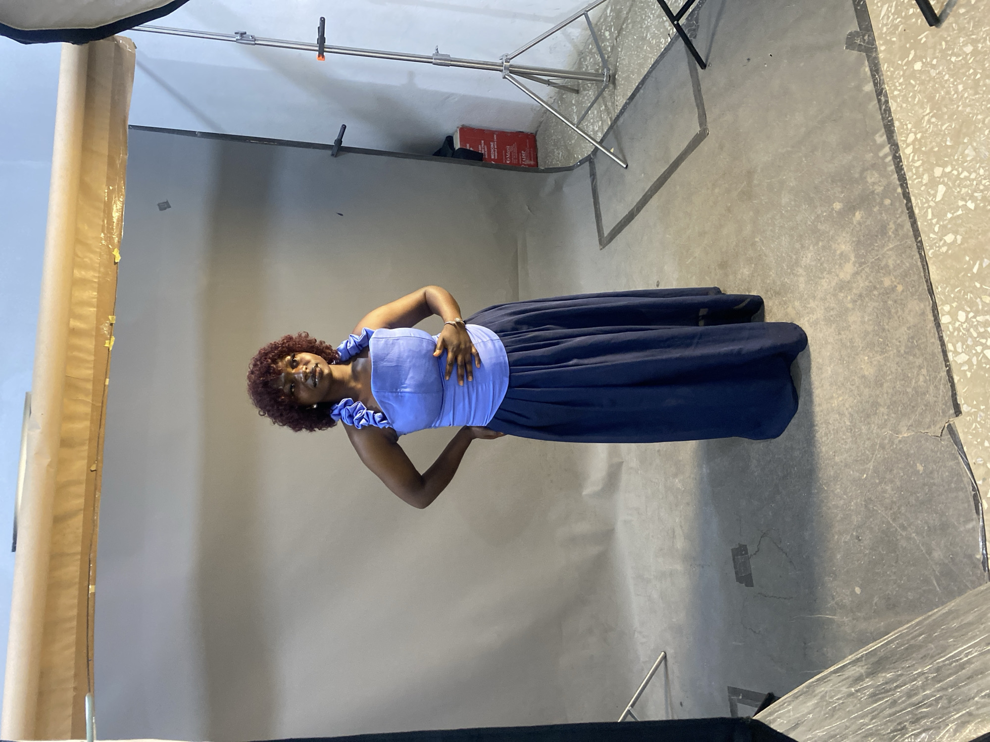 A medium shot photograph shows a woman with curly, dark hair wearing a light blue, sleeveless top with ruffled straps and a long, dark blue skirt. She stands in a studio with a gray backdrop and a concrete floor. The woman is positioned slightly off-center to the left, with her right hand on her hip and her left hand resting on her abdomen. Her head is tilted slightly upwards and to the right, and she appears to be looking towards the viewer with a relaxed expression.
In the background, professional photography equipment is visible, including a large softbox on the left and a light stand with a boom arm on the right. A roll of brown paper or fabric stands vertically against the backdrop on the left. The setting suggests a professional photo shoot. The floor has a mottled gray appearance with some lighter, speckled areas. The lighting is soft and diffused, characteristic of a studio environment. No specific time of day or weather conditions can be determined from the image. The overall mood is calm and posed. There is some visible text on a red object in the background, but it is illegible.