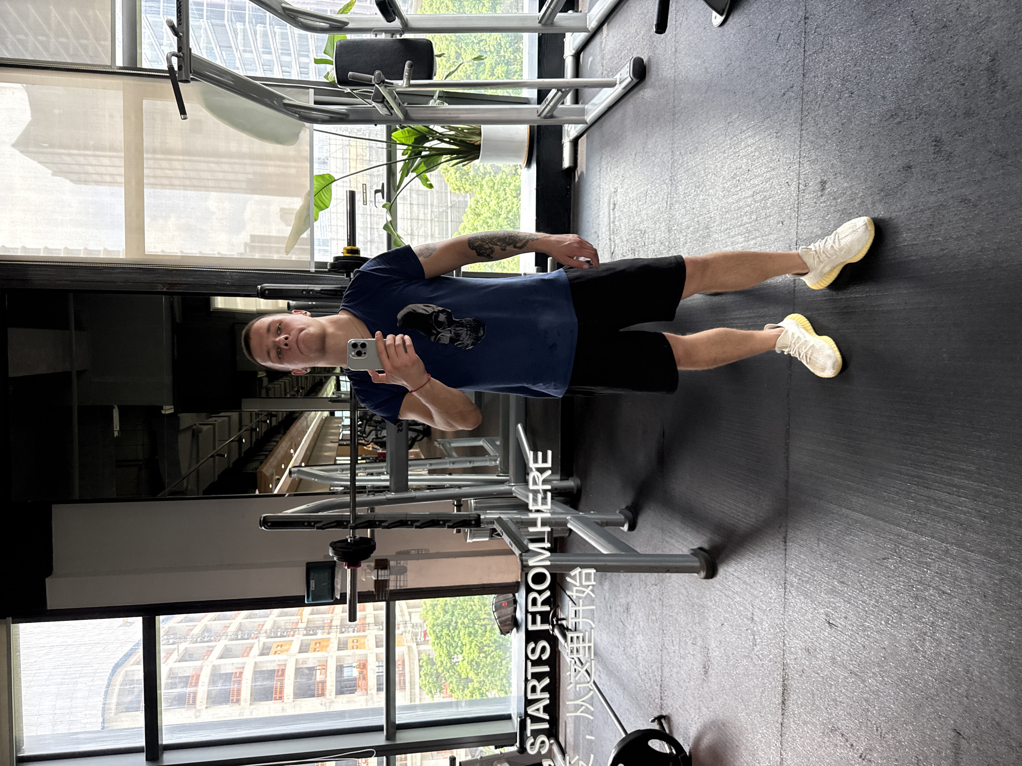 In Haizhu District, China, a man is taking a selfie in a gym. He is standing in front of a large window that offers a view of city buildings. The man is wearing a dark blue t-shirt with a skull graphic and black shorts, and white athletic shoes. He is holding a smartphone in his right hand. In the background, gym equipment, including a weight rack and exercise machines, are visible. The floor of the gym is dark and has visible lines. A large plant in a white pot is situated near the window. White text reads "STARTS FROM HERE" in English and Chinese characters, "从这里开始", positioned in the lower left quadrant of the image.