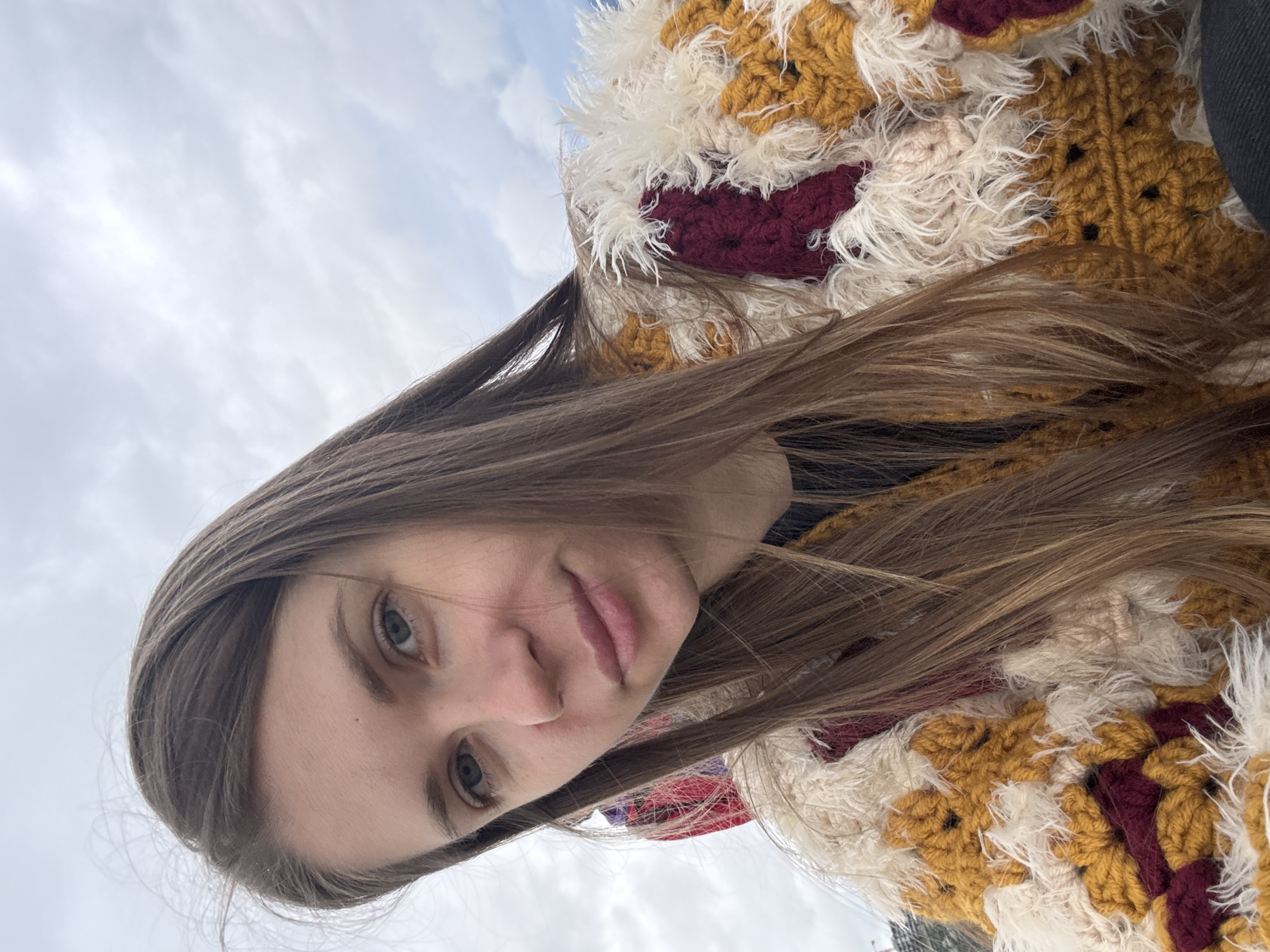 A young woman with long, brown hair is the focus of a selfie-style image taken outdoors under a cloudy sky. The woman has light skin, light-colored eyes, and is wearing a garment made of crocheted squares in various colors, including mustard yellow, cream, and maroon. The garment has fringed edges. The photograph appears to be taken from a low angle, looking up at the subject. The location is potentially Batumi, Georgia. No specific landmarks are visible.