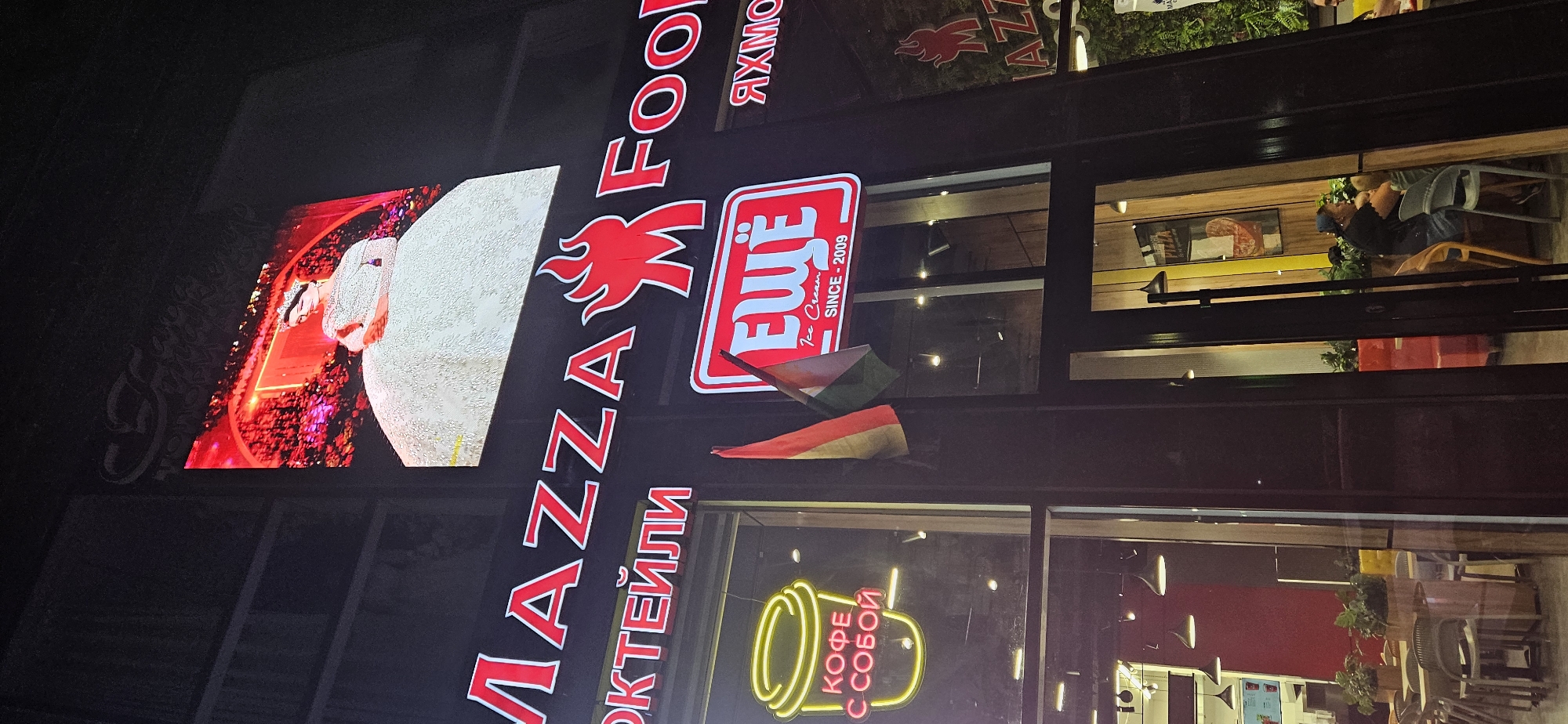 The image shows a night view of a building facade with illuminated signs and a large screen displaying an advertisement. The signs visible are "MAZZA FOOD" in large red letters with a flame logo, and a smaller red sign with white text that reads "ЕЩЁ" (which translates to "More" or "Even More") above "Ice Cream SINCE - 2009". To the left, a neon sign of a coffee cup with Russian text "КОФЕ С СОБОЙ" (Coffee To Go) is displayed in yellow. A Hungarian flag is seen hanging on the building. The large screen at the top displays an image of a woman in a white wedding dress wearing a crown. Through the glass windows of the building, interior seating areas with tables and chairs, as well as plants, are visible. People are seated inside the establishment, some of whom appear to be eating. One person in the interior is looking towards the camera. The overall scene suggests a commercial establishment, possibly a restaurant or cafe, in the city of Dushanbe, Tajikistan, given the context of the provided location. The lighting is predominantly artificial, with bright neon and LED displays illuminating the facade.