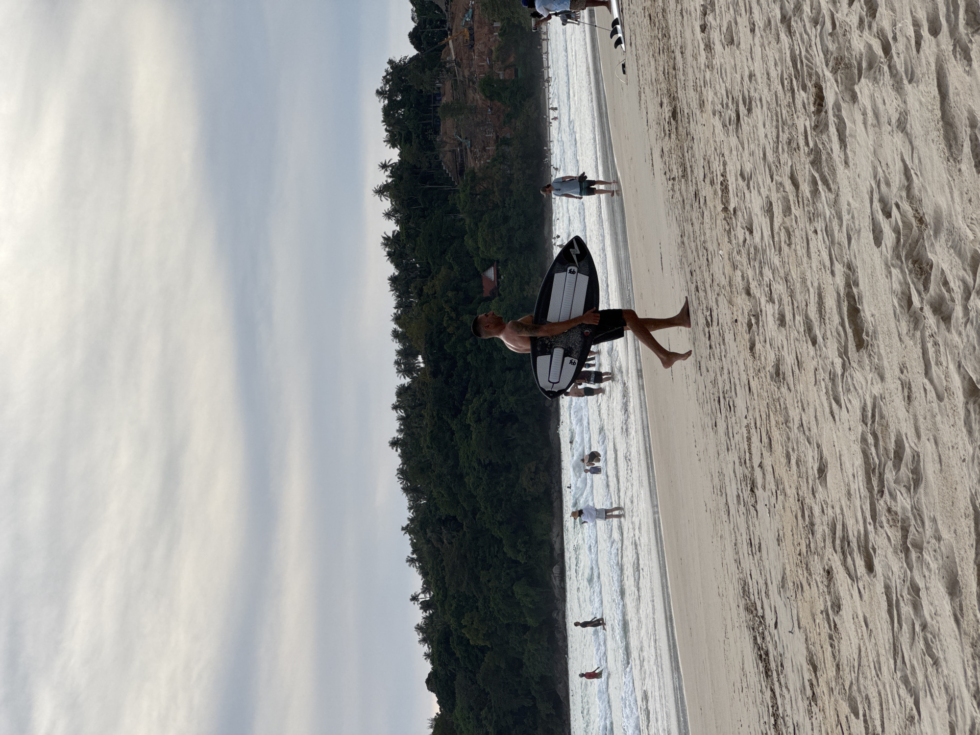 In Karon, Thailand, a man carries a surfboard on a sandy beach. The man, wearing black shorts, walks from left to right in the frame, his torso exposed. Several people are visible in the ocean behind him, some wading in the water and others swimming. The beach is lined with a dense forest of palm trees and other vegetation, with a cloudy sky overhead.