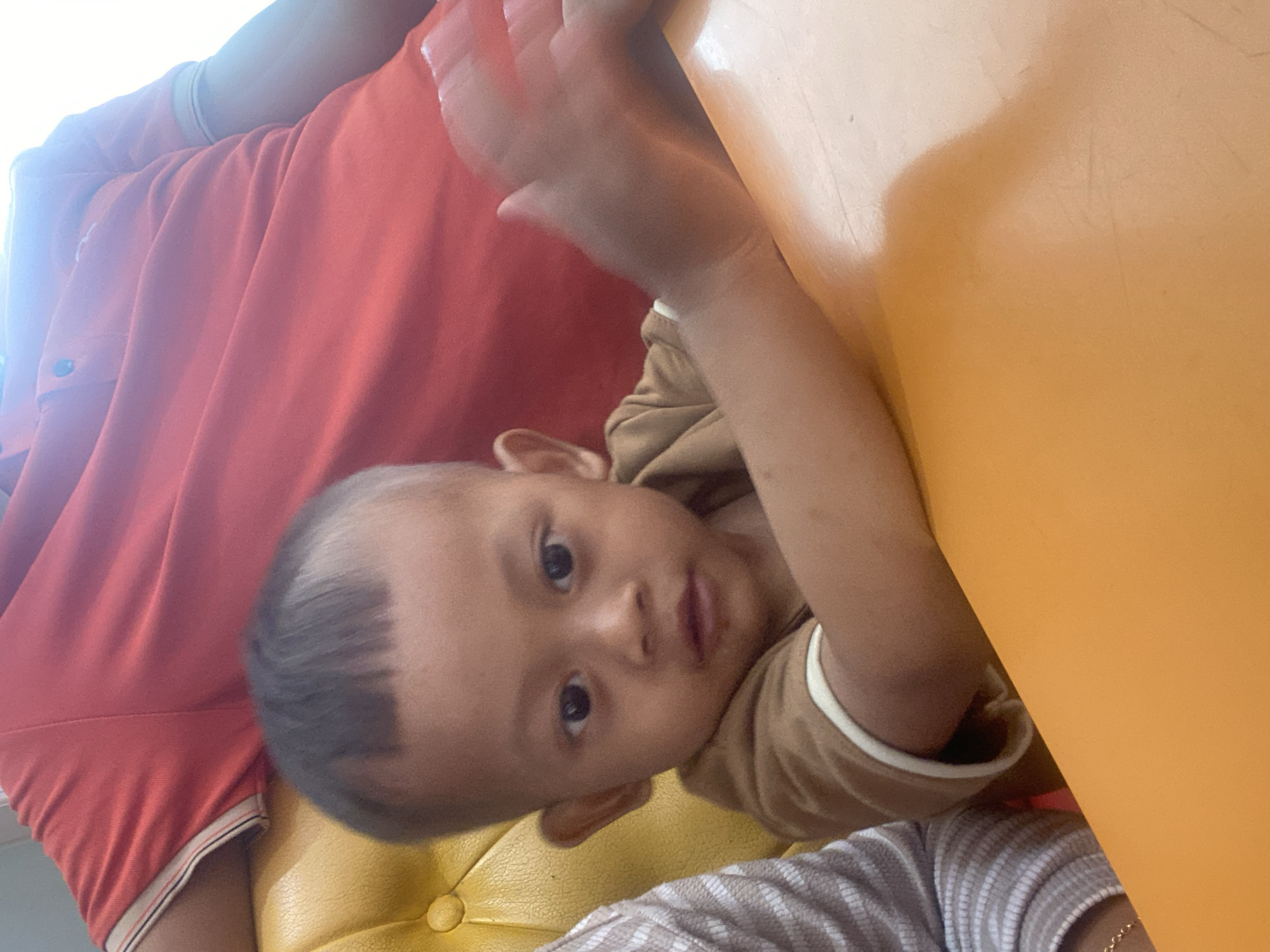 A young child with short, dark hair, wearing a brown shirt, sits at an orange table. The child is looking towards the camera, with their right hand blurred due to motion. In the background, a person in a red shirt is partially visible, seated on a yellow chair. The scene appears to be indoors, possibly a restaurant or home setting. There is no identifiable information about the specific location within Kudus, Indonesia.