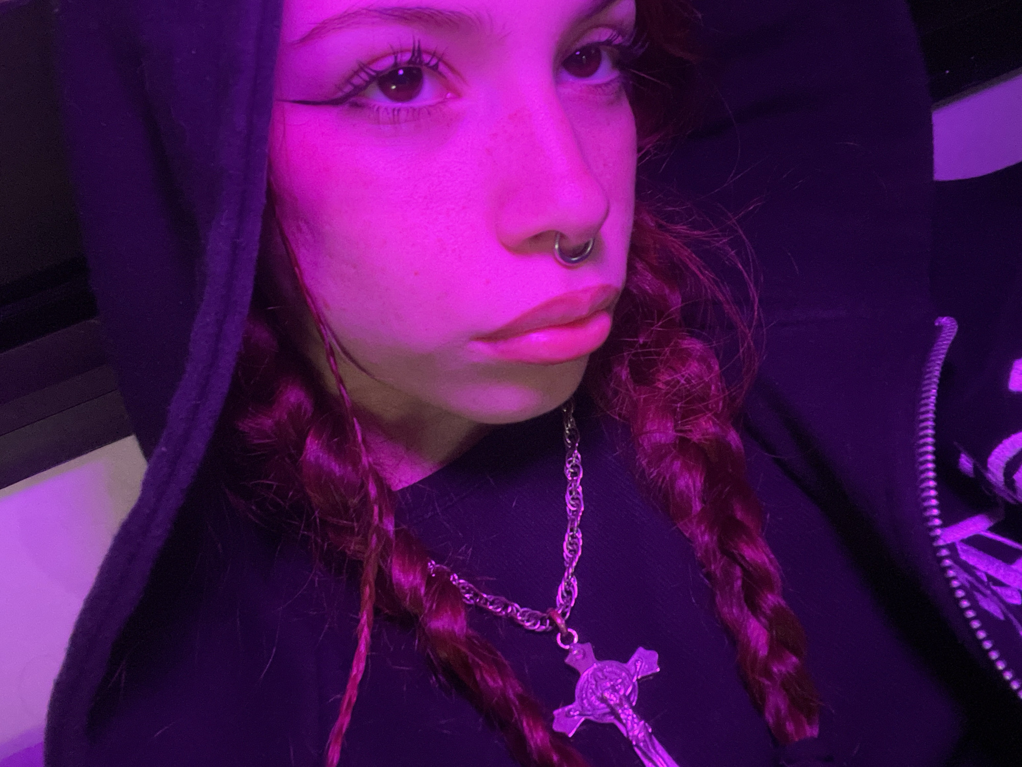A young person with light skin is wearing a black hooded sweatshirt and a cross necklace. They have reddish-brown hair in braids, and a septum piercing in their nose. The lighting is purple and pink. There is no indication of the image's location, nor any elements suggesting it was taken in Buenos Aires, Argentina.