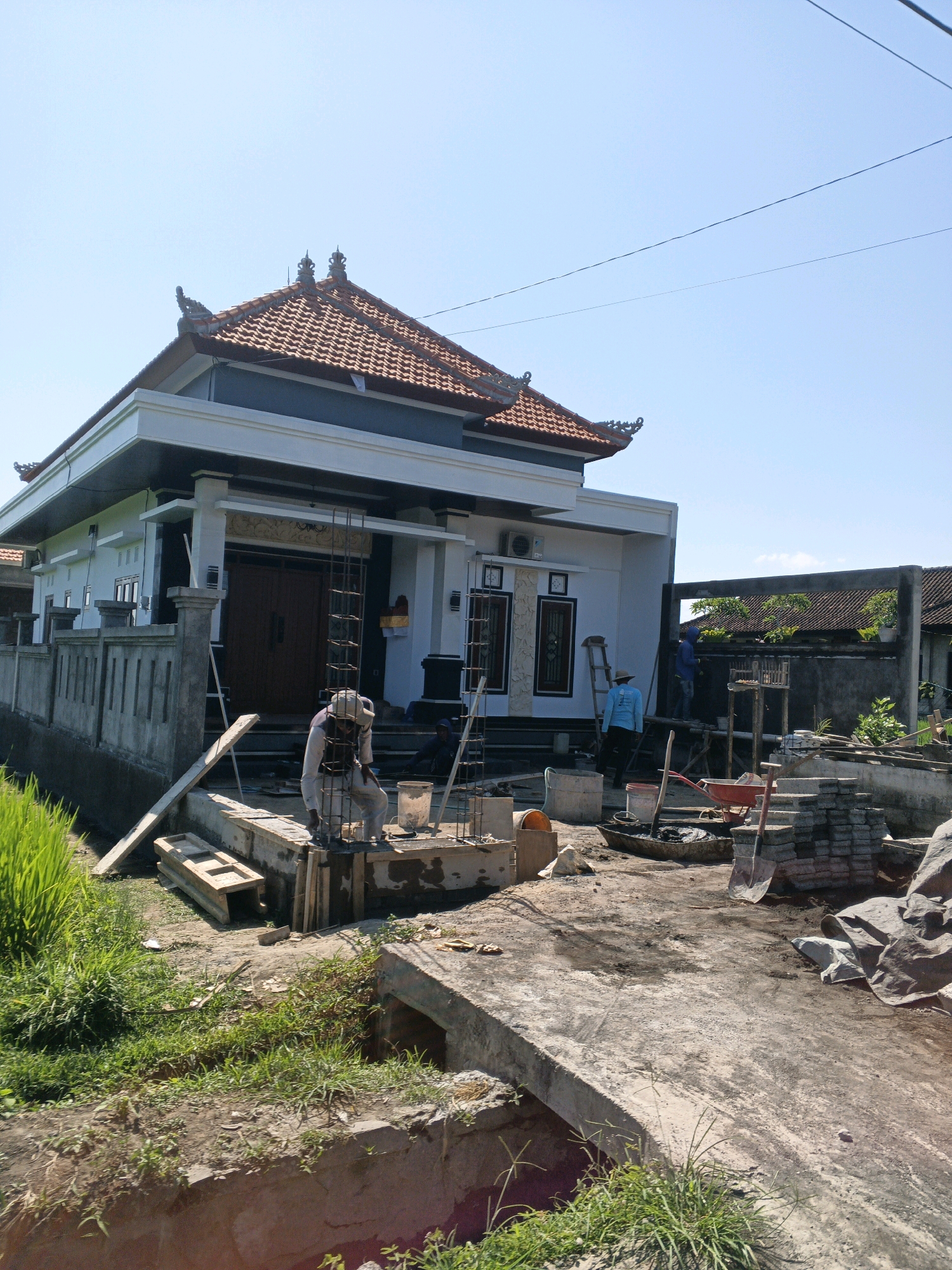 A construction scene unfolds under a clear blue sky in Abiansemal, Indonesia. A modern house with a distinctive tiered, tiled roof, characteristic of Balinese architecture, is under construction. The white facade of the house is accented with dark trim and large wooden doors. In the foreground, a construction worker, dressed in light-colored workwear and a head covering, is actively engaged in laying cement blocks and working with rebar. Other workers are visible in the background, one in a blue hoodie operating a ladder against the house, and another further back near a building. The site is littered with construction materials, including piles of bricks, a red wheelbarrow, buckets, shovels, and various wooden planks. A green rice paddy borders the construction area to the left, with a concrete structure, possibly an irrigation channel, running through the foreground. The overall impression is one of ongoing development in a semi-rural setting.