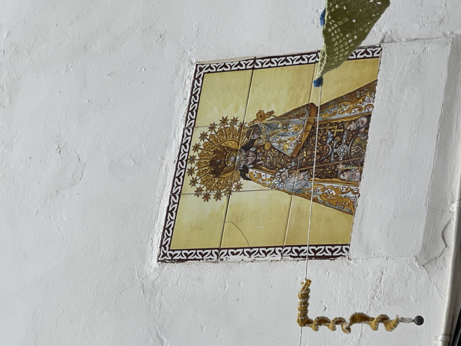 The image captures a weathered white stucco building wall in Buñol, Spain, featuring a rectangular ceramic tile panel embedded in its surface. The panel depicts a crowned female figure, likely the Virgin Mary, holding a smaller crowned figure, presumably the infant Jesus. Both figures are adorned in richly detailed robes with gold, brown, white, and blue accents. A prominent golden sunburst halo with radiating points encircles the main figure's head, set against a light yellow background. The tile panel is framed by a repeating dark brown patterned border. Multiple vertical and horizontal cracks are visible across several tiles within the panel. Two thin horizontal wires, likely clotheslines, stretch across the lower portion of the scene in front of the tile panel. From the left wire hangs a coiled, yellow-beige knitted or crocheted ornament, terminating in a small dark sphere. A section of a green, loosely woven or crocheted fabric item, possibly a decorative flag or garment, hangs from the right wire, partially visible on the right edge of the frame. The white wall exhibits minor cracks and texture variations, indicating its age and exposure. No individuals are present in the scene.