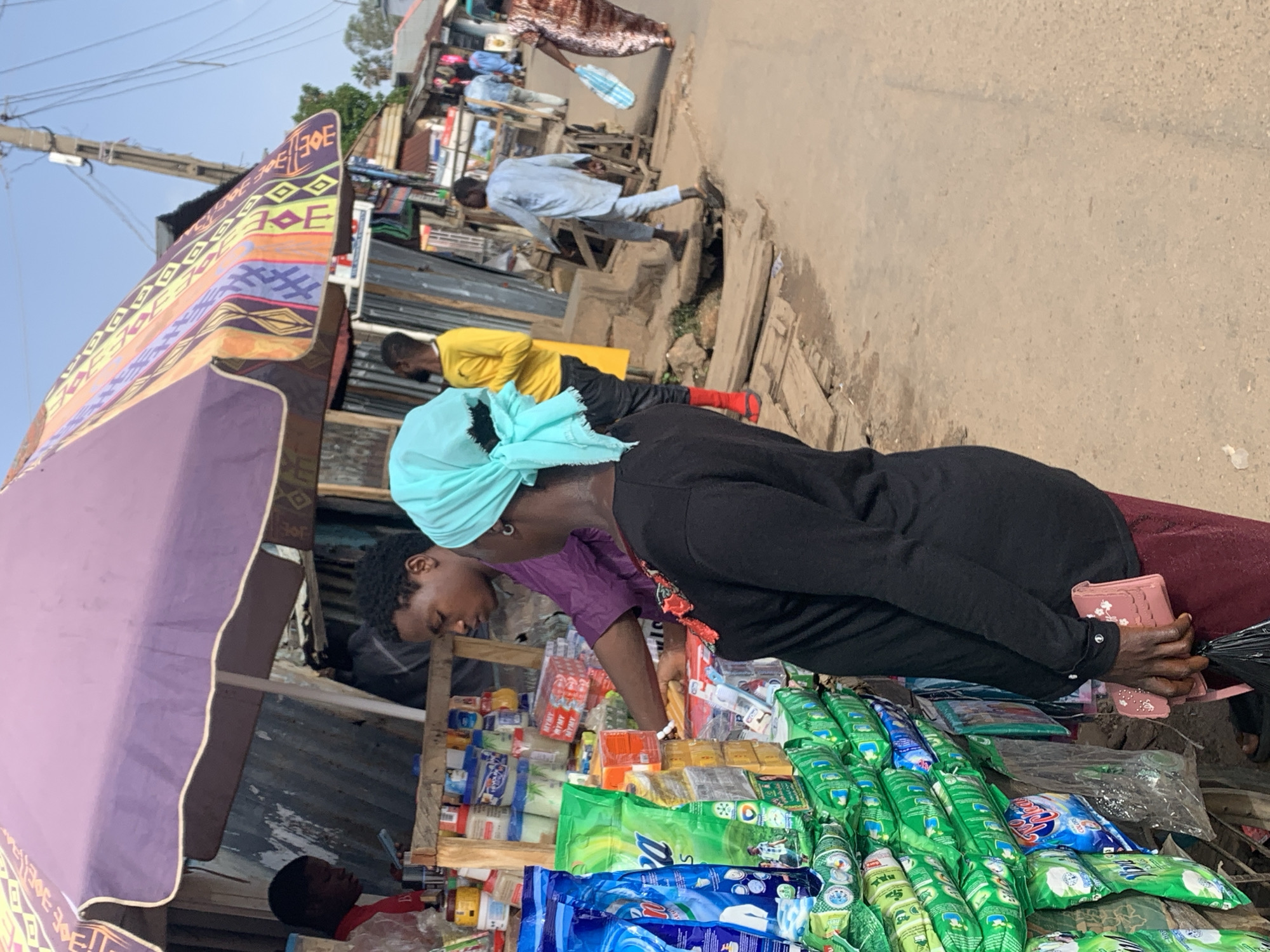The image depicts a street market scene in Jos, Nigeria. In the foreground, a woman wearing a black long-sleeved shirt and a turquoise headscarf stands facing a stall laden with green and blue packaged goods, likely detergent or cleaning supplies. She holds a pink purse with floral embellishments. A vendor, a young man in a purple shirt, is positioned behind the stall, interacting with the woman.
Behind the stall, other individuals are visible. A young man in a yellow shirt stands near the back of the stall. Further down the street, a man in a light blue outfit walks away from the camera, and a woman in a patterned dress is seen carrying a light blue bag. The market area appears to be an open-air setting with various stalls and structures lining a dirt road. The overall atmosphere suggests a bustling commercial environment.