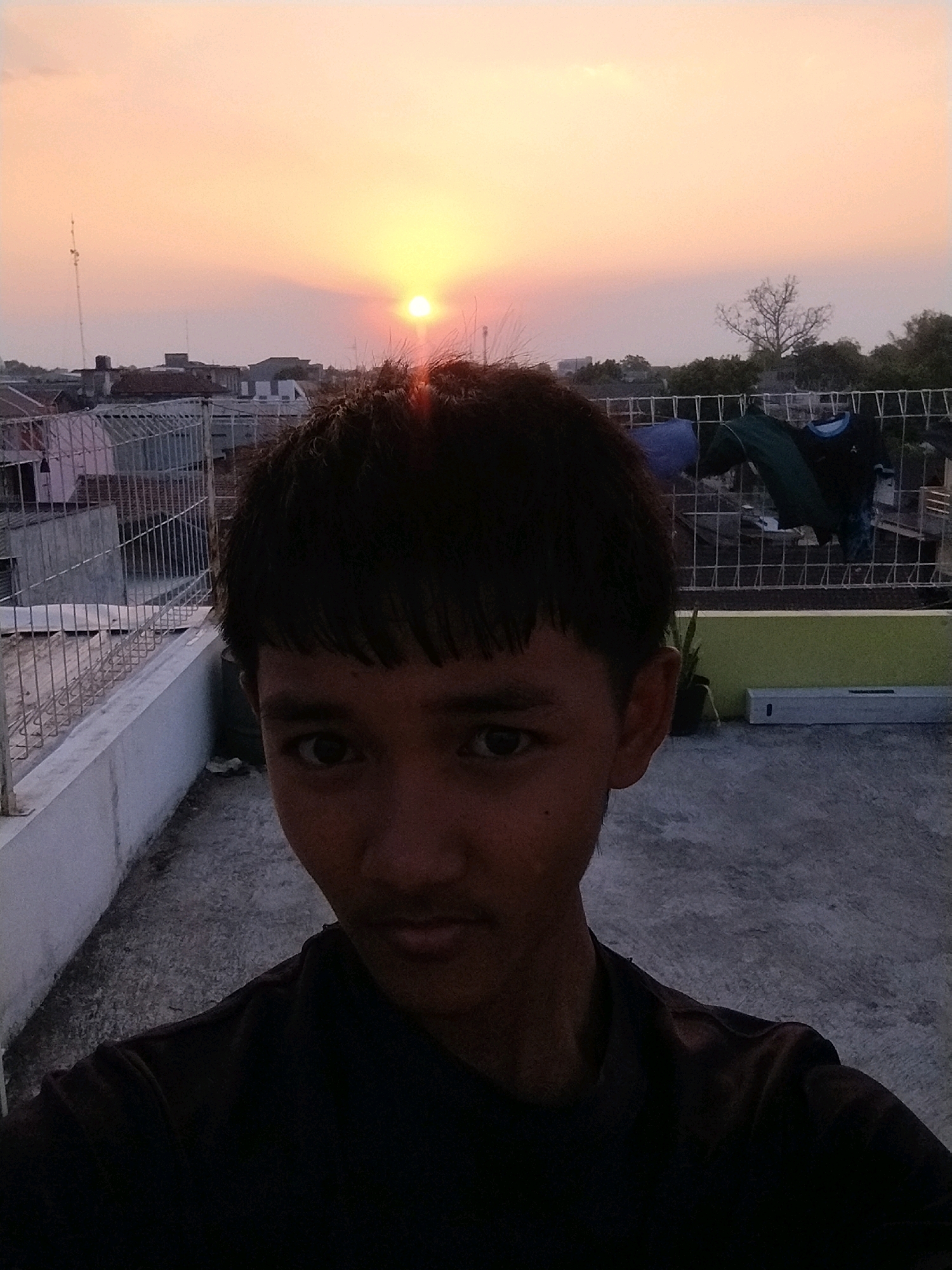 This is a selfie taken by a young male on a rooftop in Gumpang, Indonesia, during sunset. The sky is a gradient of orange and yellow, with the sun just above the horizon. In the background, there are low-rise buildings and a fence with clothes hanging on it. The foreground is dominated by the young male's face, with his dark hair falling over his forehead and his eyes looking directly at the camera. He is wearing a dark t-shirt. The rooftop is paved with concrete, and there is a white railing along the edge. The overall scene suggests a casual, everyday moment captured during a beautiful sunset.