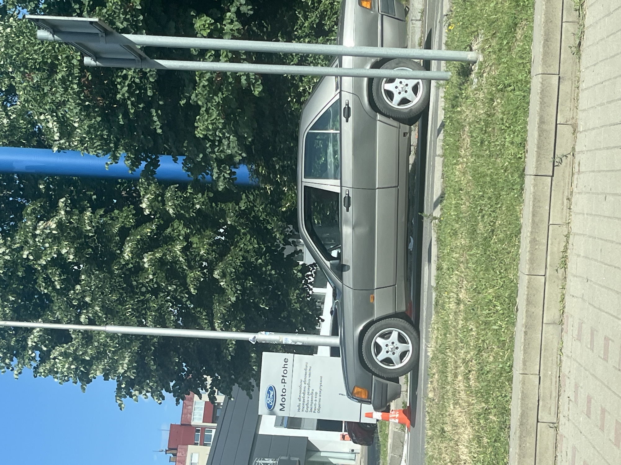 The image shows a gray Mercedes-Benz W201 sedan parked on a street in Sofia, Bulgaria. The car is in front of a Ford dealership with a sign that reads "Ford Moto-Pfohe". The dealership appears to be on a sunny day, with green trees and a blue sky in the background. A traffic cone is visible in front of the car, and a street sign is to the right of the car. The street is paved with concrete, and there is a patch of grass on the right side of the image.