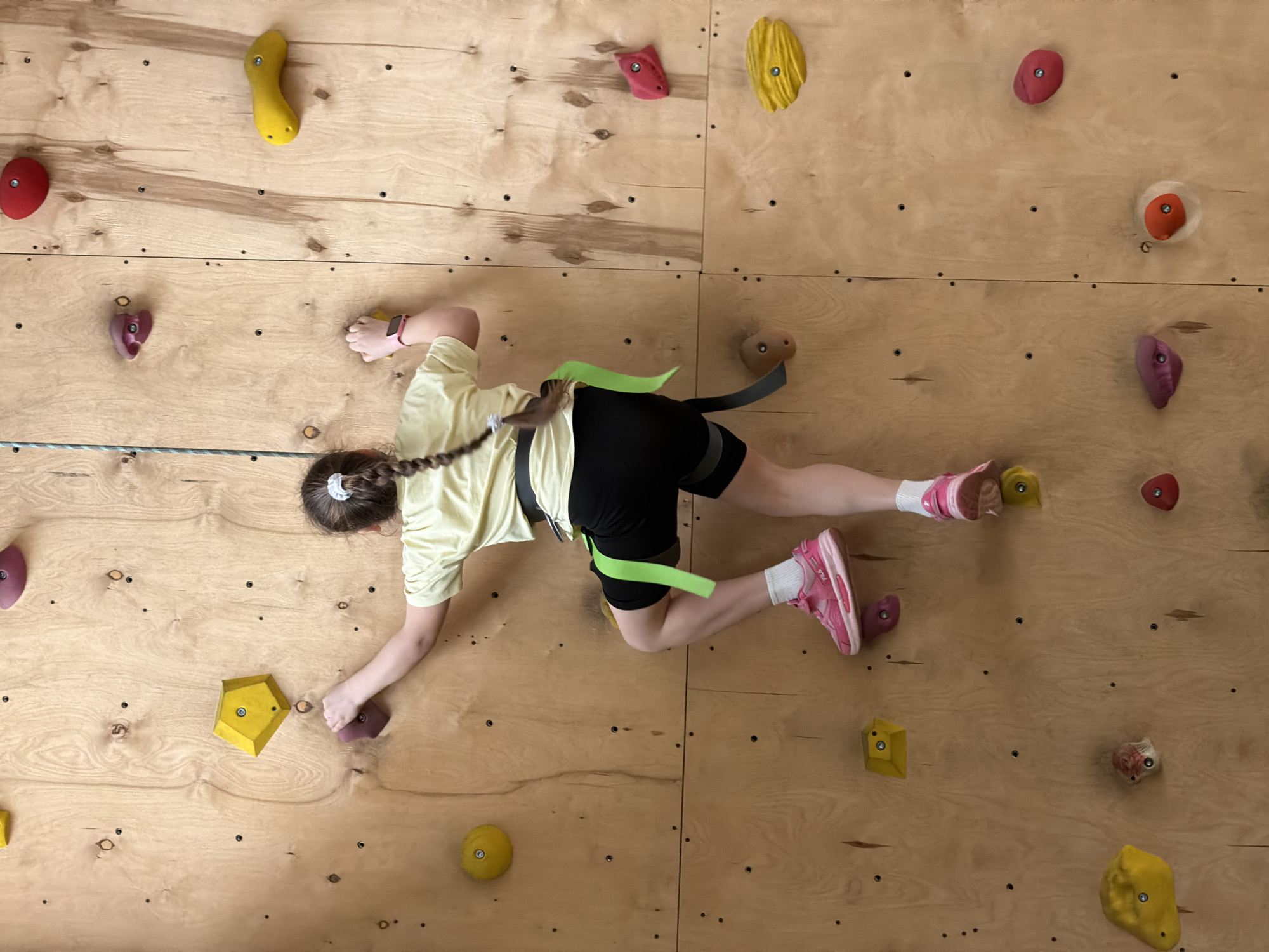 An overhead or high-angle shot captures a young person actively climbing on a wooden bouldering wall in an indoor climbing gym, located in Izhevsk, Russia.
The climber, a child or teenager with brown hair tied in a braid, is dressed in a light yellow short-sleeved t-shirt, black shorts, white socks, and pink sneakers. A black climbing harness with a distinctive neon green loop is secured around their waist and thighs. The person is mid-climb, demonstrating a dynamic stretch with their left hand reaching for a purple hold and their right foot firmly planted on a yellow hold. Their body is horizontally oriented across the wall, showcasing a moment of concentration and physical exertion.
The climbing wall is constructed from light-toned wooden panels, fitted with numerous small black bolts for attaching a variety of brightly colored climbing holds in yellow, red, purple, and beige tones. A metallic cable or rope is visible horizontally across the lower-left portion of the frame, likely part of a safety or support system. Visible details include a pink wristband on the climber's left arm and the partial branding "FILA" on their pink sneakers.