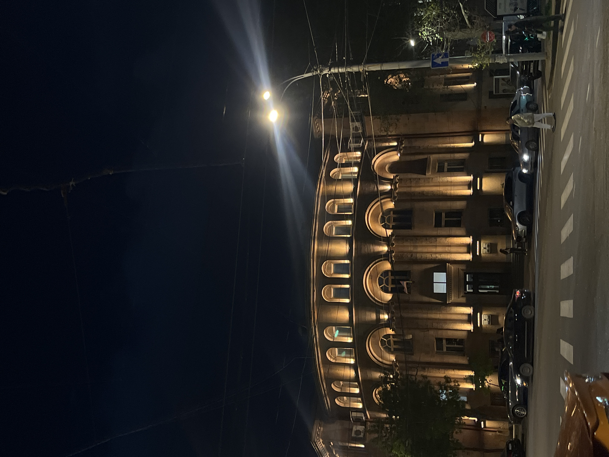 The image, originally rotated 90 degrees clockwise, depicts a lively urban street scene at night. The prominent subject is a grand, multi-story building with a curved facade, illuminated by warm, upward-shining spotlights. Its classical architecture features numerous arched and rectangular windows, with intricate details highlighted by the artificial light.
On the street level, a pedestrian crossing with white zebra stripes is visible. A person wearing light-colored clothing (jacket and trousers) is walking across this crossing. Several vehicles populate the street: a light blue sedan is near the crosswalk, a darker car is parked behind it, and the front of a golden-yellow car is partially visible in the bottom right corner.
A complex network of overhead utility wires stretches across the dark night sky, supported by poles. Bright streetlights, attached to these poles, emit strong beams and noticeable lens flares. One pole near the pedestrian crossing displays two traffic signs: a blue rectangular sign with a white arrow (pointing left if the image were upright) and a red circular sign with a horizontal white bar, typically indicating "no entry." Trees with sparse foliage are also present along the street.
The scene captures a typical evening in an urban setting, likely Yerevan, Armenia, characterized by extensive artificial lighting that defines the architecture and street activity. No other legible text is visible on buildings or signs.