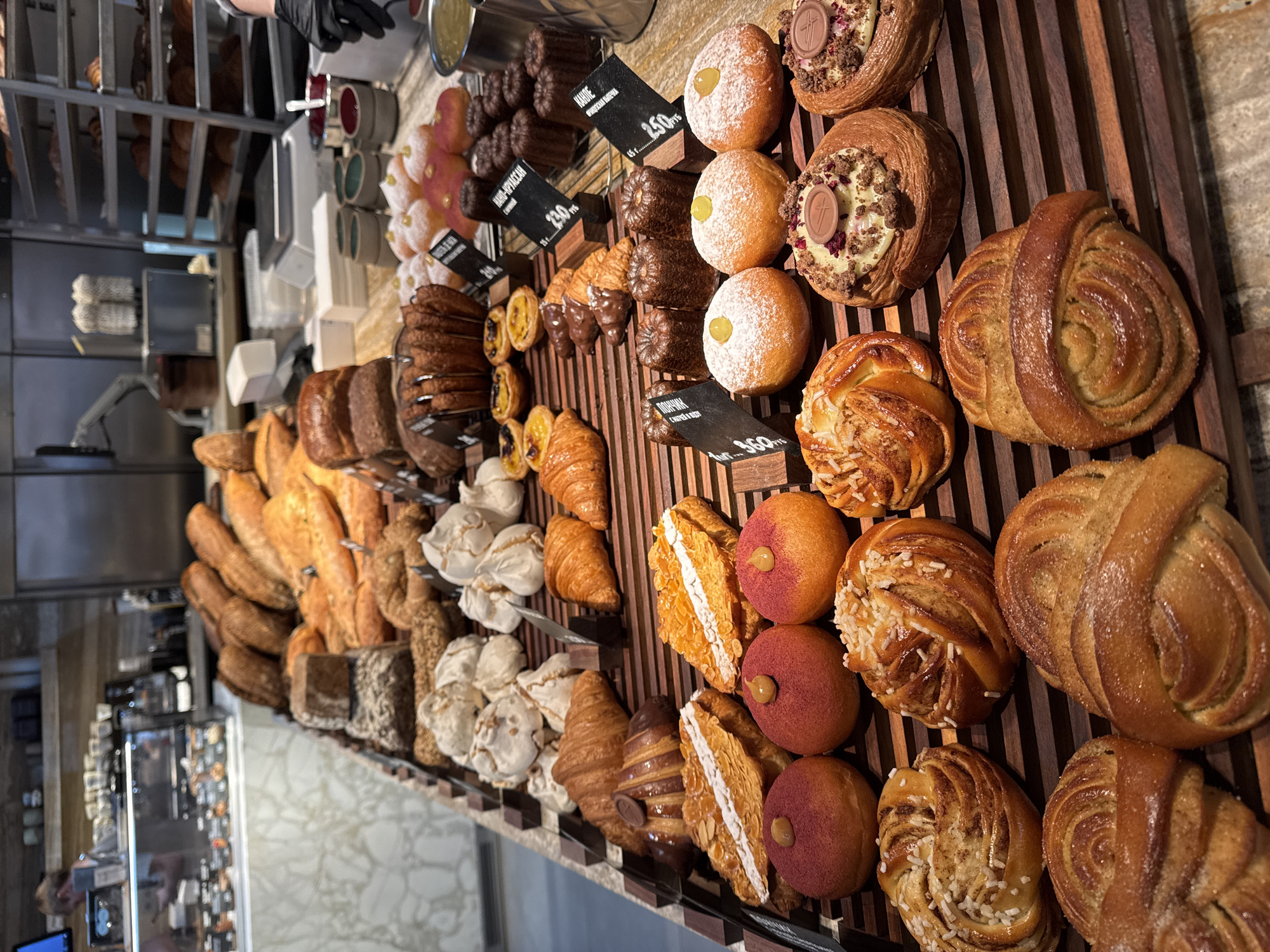 A wide-angle, overhead shot captures a bakery display. The display features rows of various pastries including breads, croissants, meringue, canelé, and filled doughnuts. Small, black price tags are placed near some of the baked goods. A gloved hand is visible near the top left corner, suggesting a person is behind the counter. Shelves stacked with baked goods can be seen in the background. The location is an unknown bakery, at an unknown location.
