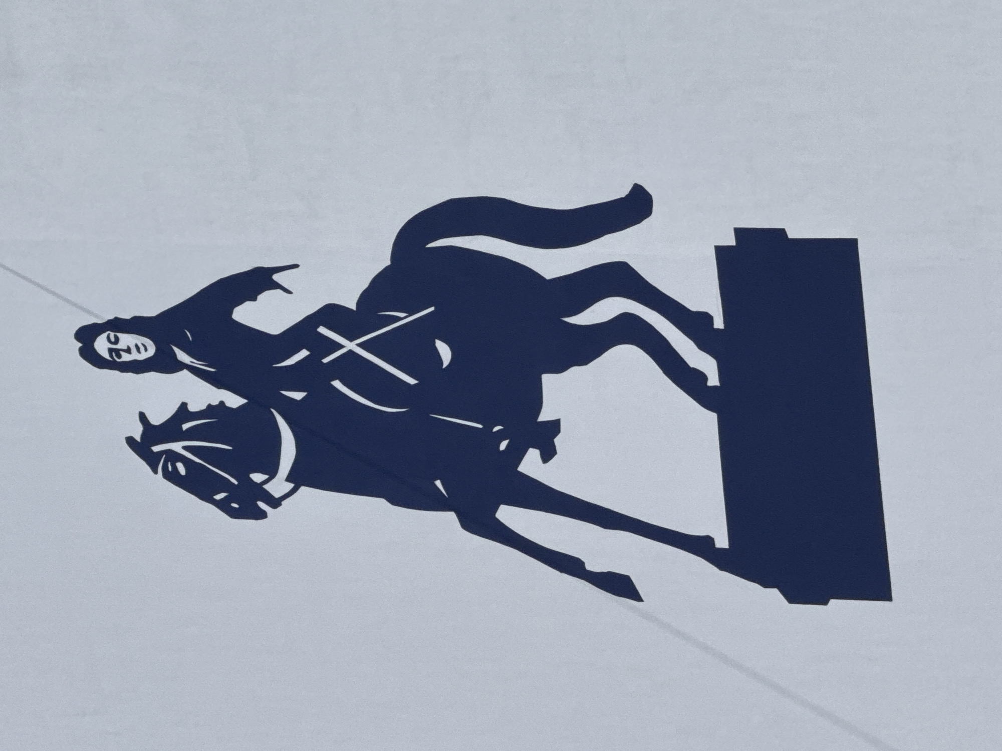 A two-tone, stylized graphic depicts a dynamic scene of a person riding a horse. The entire image is rendered as a dark blue silhouette against a plain, light grey background, giving it a minimalist aesthetic.
The central subjects are a rider and a horse, positioned at an angle, suggesting motion from left to right and slightly upwards. The rider, likely male, is shown in profile, wearing what appears to be a hood or hat, with facial features such as an eye, nose, and mouth faintly visible. His right arm is raised and extended forward, with the index finger pointing. A prominent 'X' mark is visible on the rider's chest.
The horse is depicted in a powerful, airborne stance, with its front legs bent and its hind legs extended, as if mid-jump or rearing. Its head is detailed with an eye, nostril, and mouth. To the right of the horse and rider is a tall, rectangular dark blue object, resembling a barrel or pillar, which the horse appears to be moving past or over.
The overall activity suggests an equestrian event, possibly barrel racing, show jumping, or a similar competition requiring agility and skill. The graphic conveys a sense of speed and action. There is no visible text in the image. Faint diagonal lines are present in the light grey background, potentially indicating the texture of the surface on which the graphic is displayed.