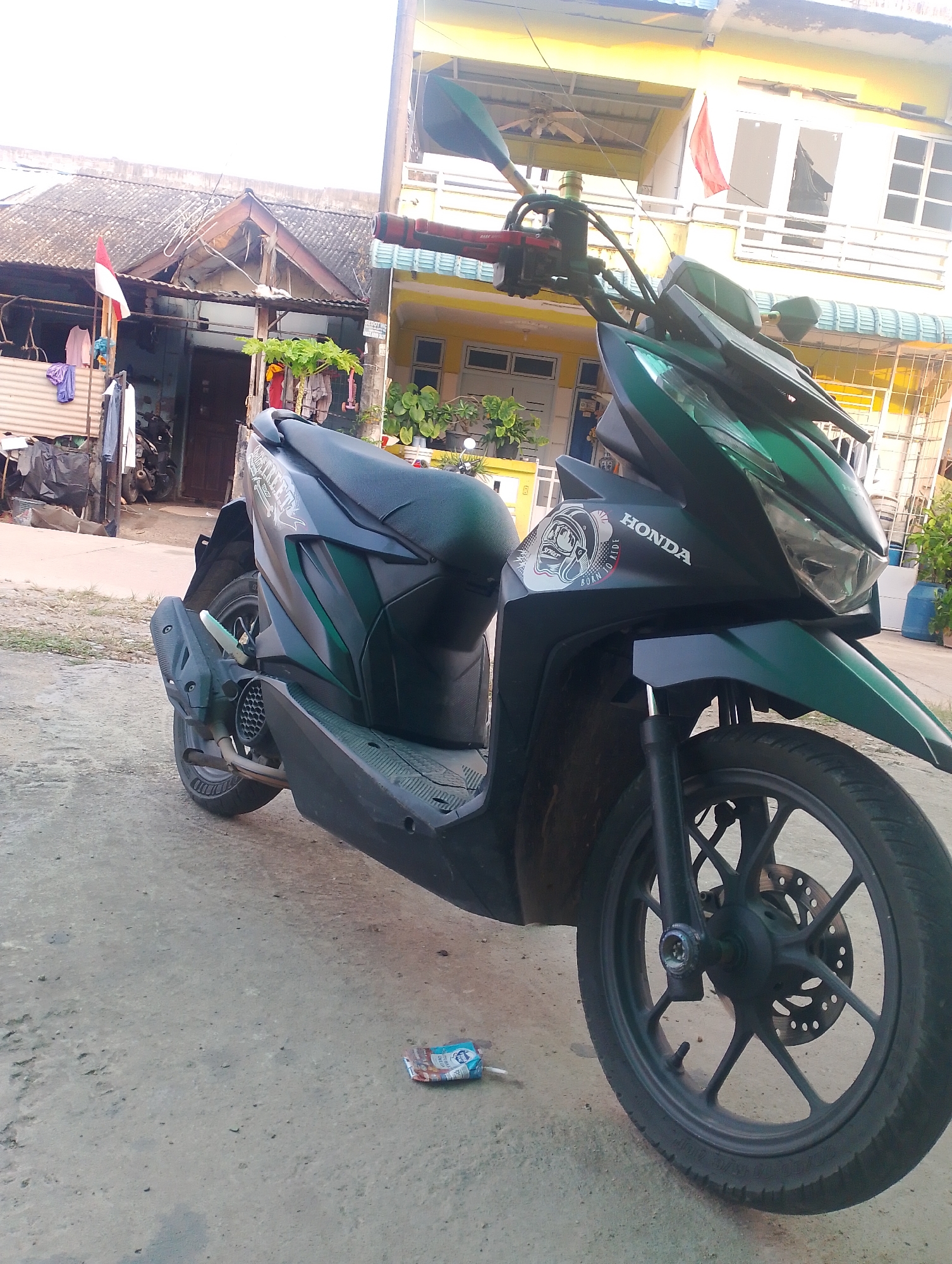 The image shows a dark green Honda scooter parked on a dirt and concrete road in front of a row of houses. The houses are painted yellow and have a weathered appearance. Some laundry is hanging on a line in front of one of the houses, and a small red and white flag is also visible. The scooter is the main subject of the image, and it is shown from the side, with the front wheel and part of the handlebars visible. The environment appears to be a residential area in Batam, Indonesia.