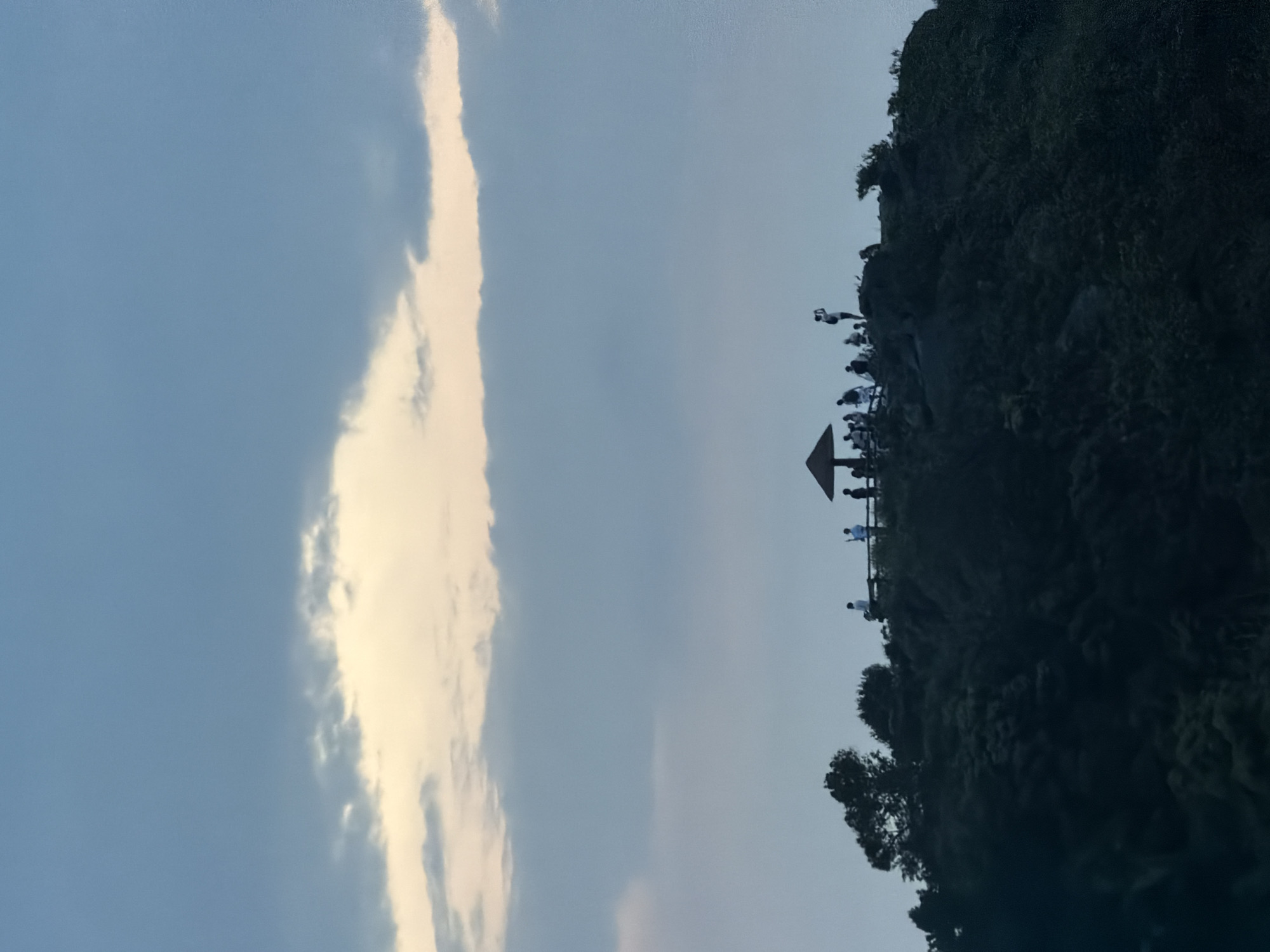 The image shows a scene on the edge of a cliff overlooking a cloudy sky. On the right, the cliff is covered with lush greenery and rocks. A viewing platform with a shelter is situated at the edge of the cliff. Many people are standing on the platform, appearing as small silhouettes against the sky. The left side of the image is dominated by a blue sky with prominent white clouds. The photograph may have been taken in the city of Đà Nẵng, Vietnam.
