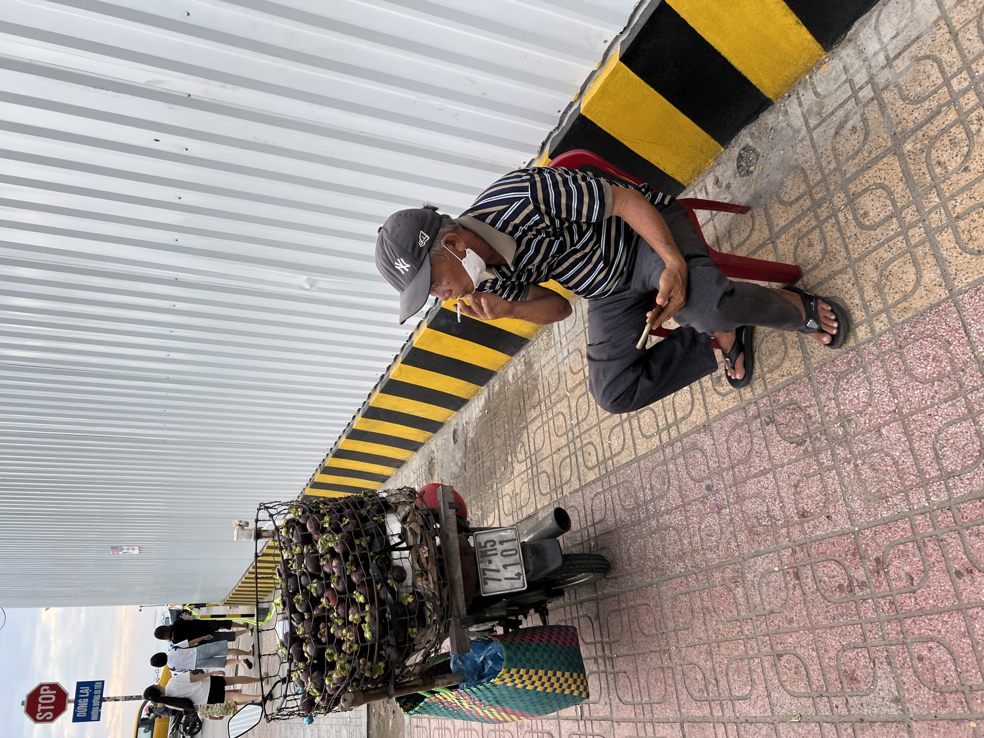 The image depicts a street scene in Nha Trang, Vietnam, with a focus on a man sitting on a red plastic chair. The man, who appears to be of Asian descent and middle-aged or older, is wearing a dark baseball cap, a striped t-shirt, dark pants, and black flip-flops. A white face mask is pulled down to his chin. He is holding a smartphone in his left hand and smoking a cigarette, with the cigarette in his right hand near his mouth.
To the left of the man, a motorcycle is parked. The motorcycle is laden with a large basket full of mangosteens, a fruit. A colorful woven bag is attached to the side of the motorcycle. The motorcycle has a license plate that reads "77-M5 4101".
The setting includes a sidewalk paved with patterned red and beige tiles. A white corrugated metal wall runs along the background, with a black and yellow striped border at its base. A "STOP" sign and a blue sign with Vietnamese text are visible on a pole to the left. Several people, including women and a child, are walking in the background, near a yellow vehicle. The sky above is overcast.