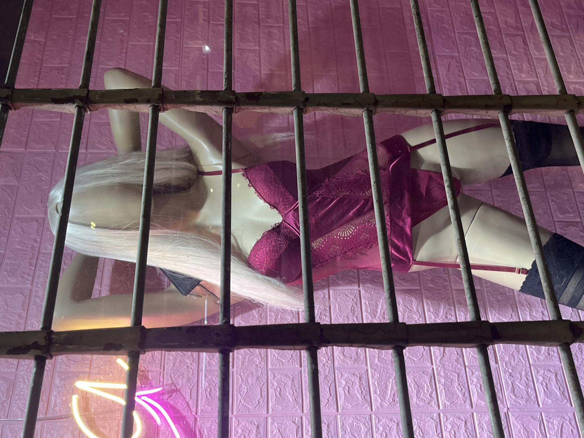 A mannequin with blonde hair is lying on a pink, textured surface behind a metal grate. The mannequin is wearing a red lingerie set and black stockings. A neon sign is visible in the lower left corner. The scene is likely a storefront window display. It is difficult to confirm the exact location is Buenos Aires, Argentina, from the image alone.