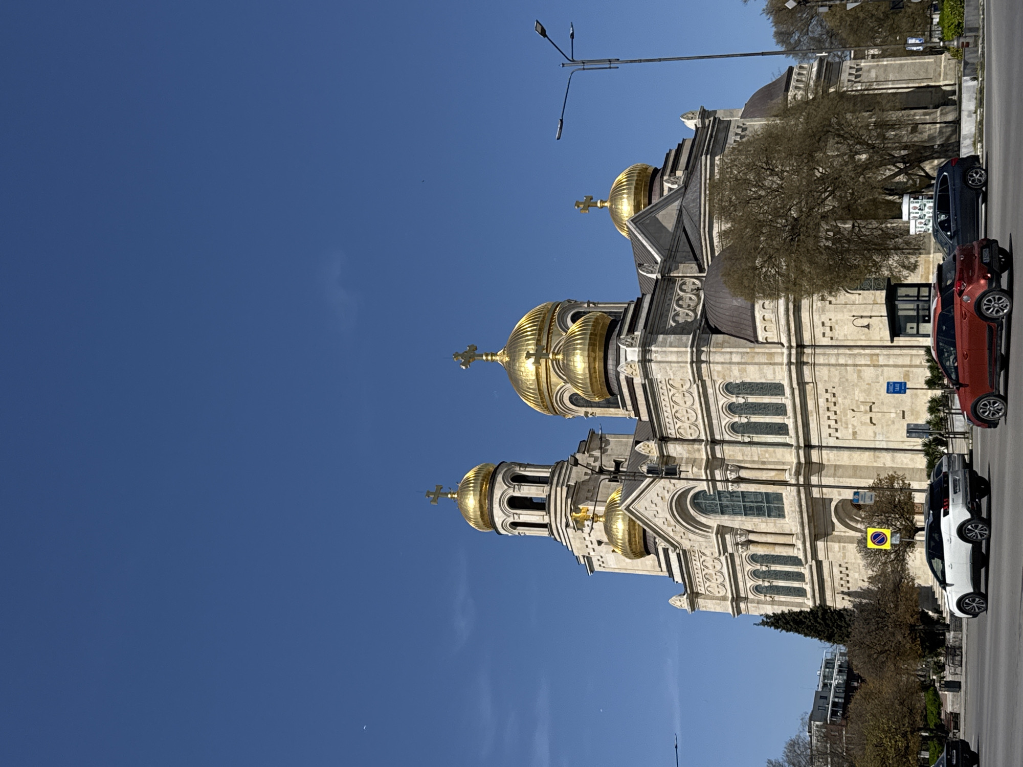 The image displays a grand Eastern Orthodox cathedral, likely the Dormition of the Theotokos Cathedral, set against a clear, bright blue sky. The cathedral features multiple gleaming golden domes, each topped with a cross, and is constructed from light-colored stone with intricate architectural details, including arched windows and decorative patterns.
The scene depicts an urban setting during the daytime, with ample sunlight illuminating the building and casting shadows. A paved street runs parallel to the cathedral, and several cars are parked along the curb, including a red car and a white SUV. Trees, some appearing bare, suggesting late autumn, winter, or early spring, are visible between the street and the building.
Notable details include a tall streetlamp extending into the sky above the cathedral. Visible street signs include a yellow circular sign with a red diagonal line across a blue background, indicating "No Stopping" or "No Parking." A partially visible blue rectangular sign that appears to say "TAXI" can also be seen above one of the parked cars. The weather is clear and sunny, enhancing the golden luster of the domes. The image is oriented vertically (rotated 90 degrees clockwise), showing the building and street rising upwards.