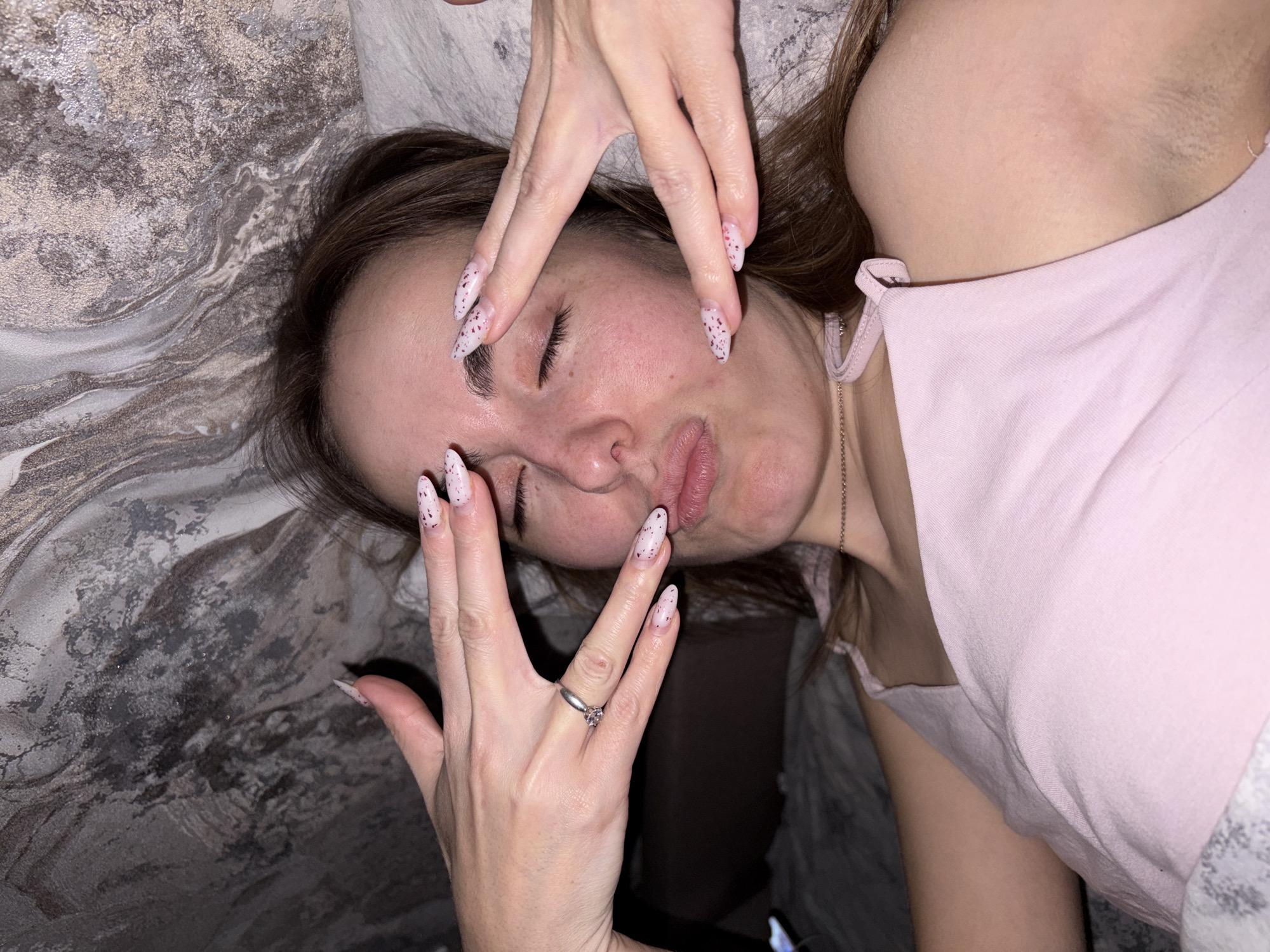 A woman with light brown hair and light skin is lying on a bed with a patterned gray comforter. She is wearing a light pink tank top and a silver ring is visible on her left hand. Her hands are positioned over her face in a triangular shape. The location of the image is not determinable from the provided image.