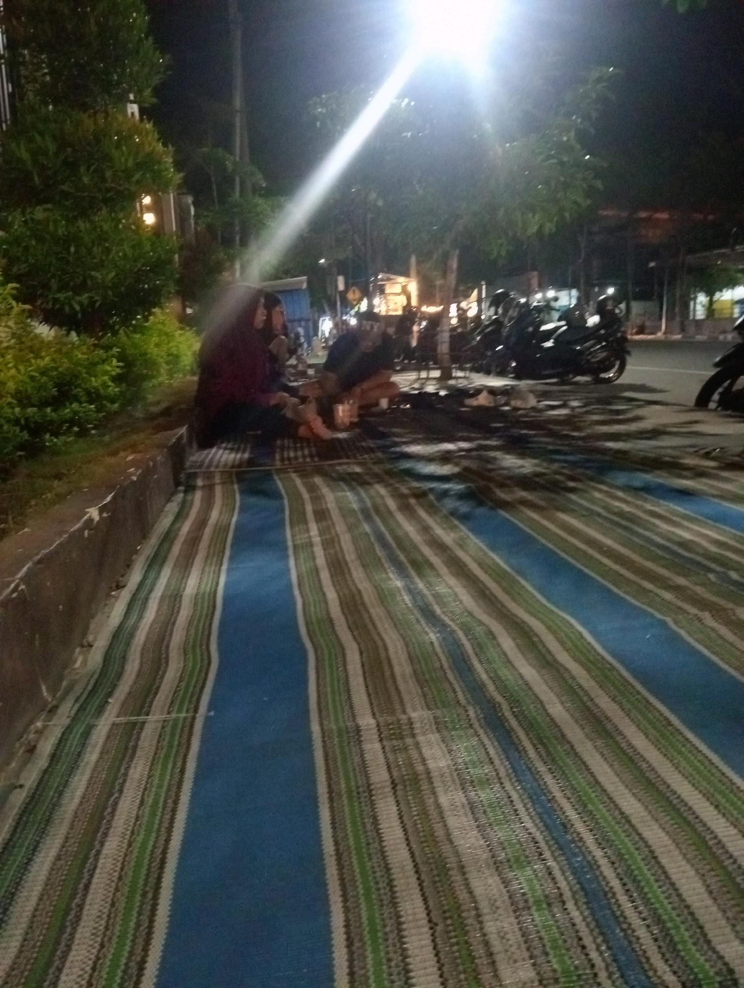 The image depicts a nighttime scene on a street in Bojonegoro, Indonesia, possibly near a row of shops or food stalls, indicated by the presence of motorcycles parked along the road and a bright street light overhead. A group of individuals are seated on a large, striped mat laid out on the ground. The mat is predominantly blue with thin, alternating stripes of green and brown. The people are casually dressed and appear to be engaged in conversation or resting. In the background, there are trees and foliage, suggesting a semi-urban or park-like setting. The lighting is dim, with the street light casting strong shadows and highlights.