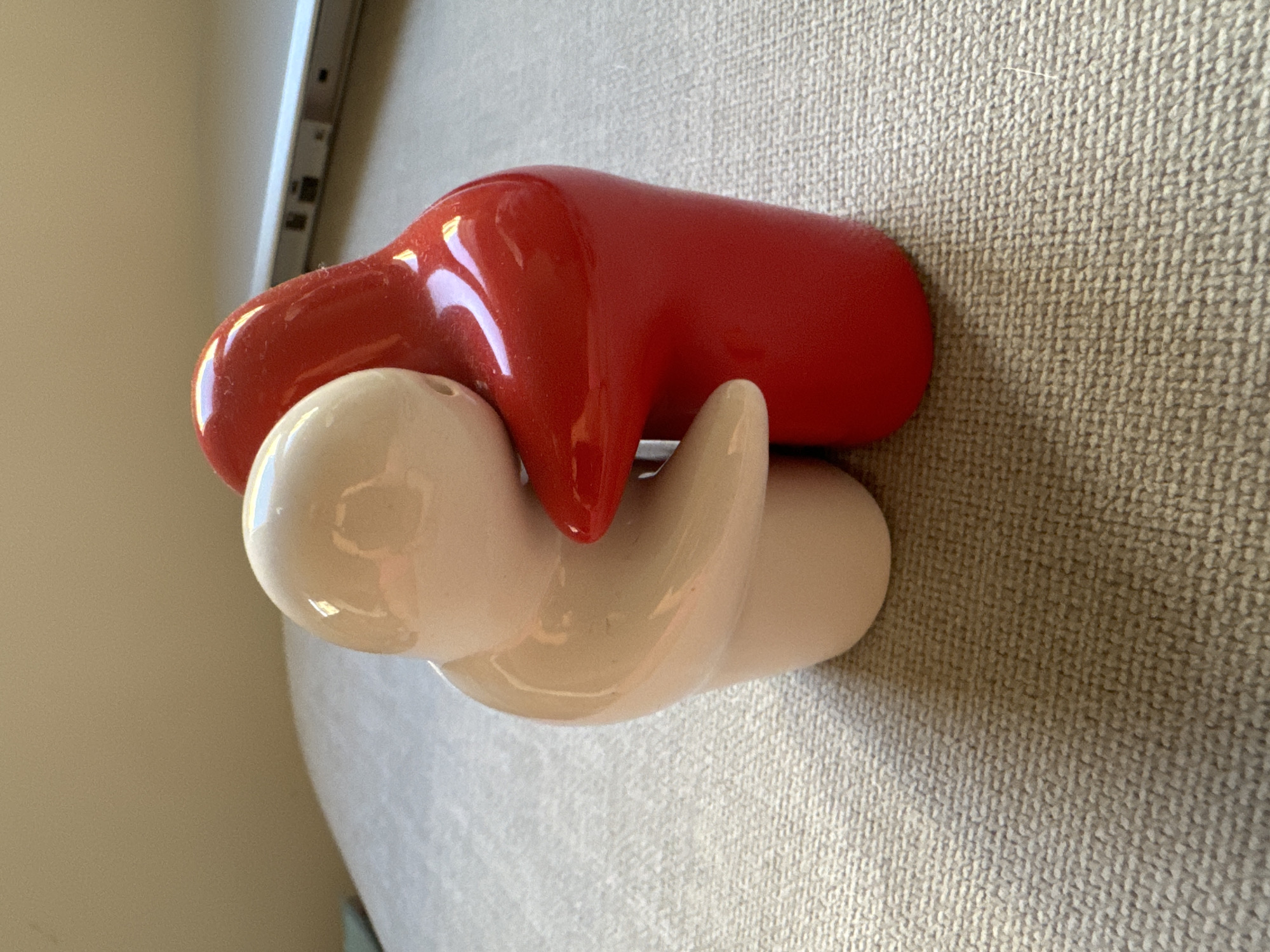 A close-up shot features two ceramic figures embracing. The figure on the left is a light beige, while the figure on the right is bright red. They are positioned on a textured, light-colored surface, possibly a woven fabric. In the background, a portion of a metallic laptop or electronic device is visible, along with a plain, light-colored wall. The lighting suggests an indoor setting with natural light. The image does not provide any information indicating a location in the city of Murcia, Spain.