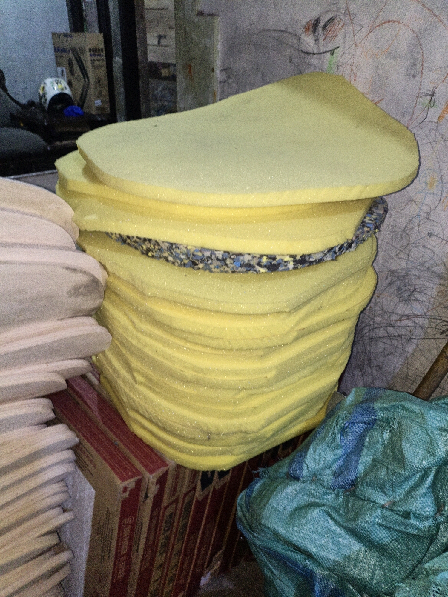 This image captures a cluttered storage area, likely in a workshop or warehouse in Cikal, Indonesia. In the foreground, a large stack of yellow foam padding is the central focus, bound by a strip of patterned material. To the left, a stack of light-colored, curved wooden objects, possibly furniture components or parts of a larger item, leans against the foam. Behind these, several cardboard boxes with red and gold labeling are visible. To the right of the foam, a large, crumpled green plastic bag is partially obscuring another object, possibly more padding or material. The background consists of a textured wall with child-like drawings and some darker vertical structures, suggesting an unfinished or utility space. The overall impression is one of a working environment with materials stored for production or repair.