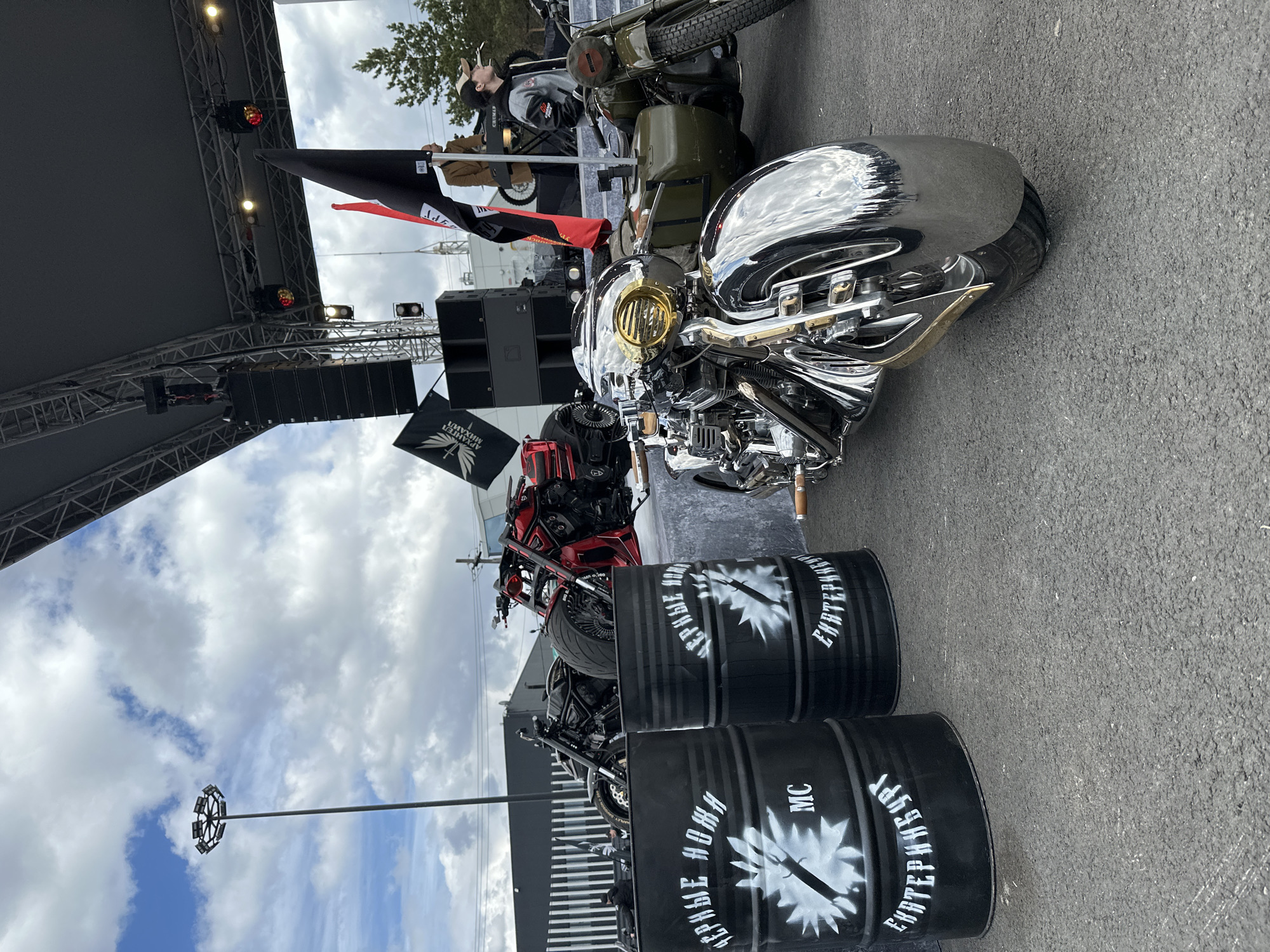The image shows a motorcycle display at an event, likely in Yekaterinburg, Russia, based on the barrel markings. The foreground features two black barrels with white stenciled text and a logo, which read "ЧЕРНЫЕ НОЖИ" (Black Knives) and "ЕКАТЕРИНБУРГ" (Yekaterinburg), followed by "MC." Behind the barrels are several motorcycles, including a chrome-plated model with a golden headlight and others in red and olive green. The background includes a stage structure with speakers and flags, one of which has the same logo as the barrels. Several people are on the stage. The sky is cloudy. A tall street light is visible in the background. The ground is asphalt.