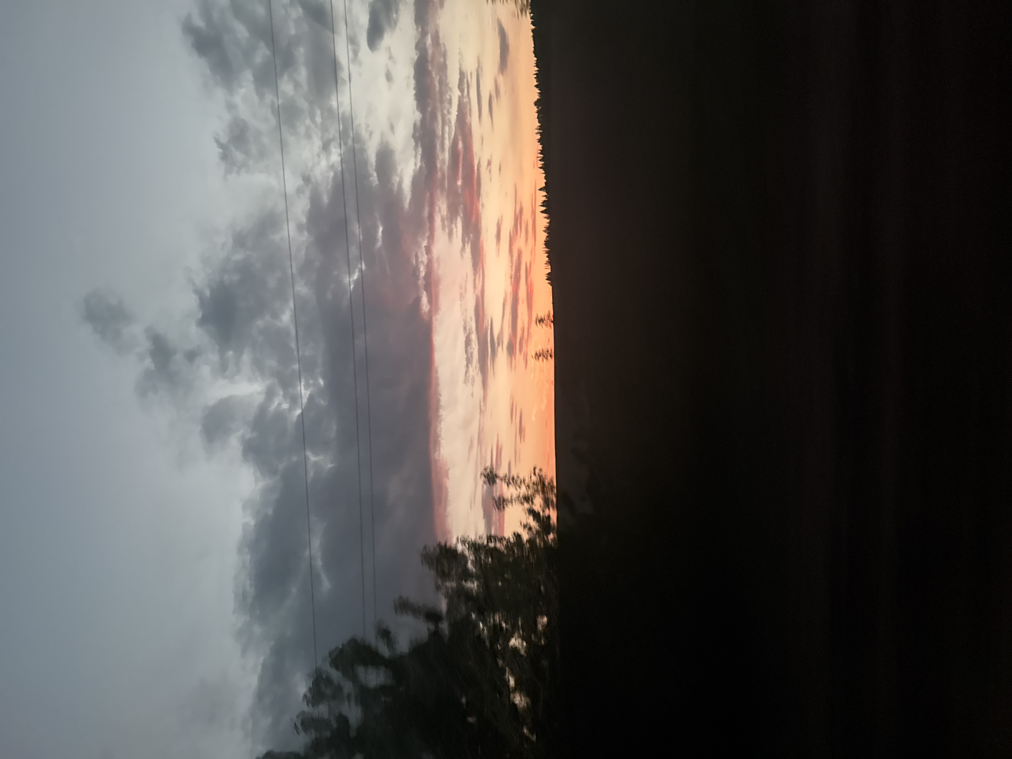 The image shows a sky with a dramatic contrast between a dark, overcast area with low-hanging clouds on the left and a sunset with orange and pink hues on the right. Thin power lines stretch across the cloudy portion. A line of trees is silhouetted against the sunset. A dark, indistinct foreground occupies the right side and bottom of the image. The setting appears to be rural due to the presence of trees and open sky.
I don't have the location information for Галево, Russia, I don't have any details about any people.